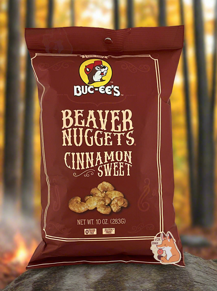A picture of the front of a opaque, brown bag of Buc-ee's Beaver Nuggets, Cinnamon Sweet.  which are puffed corn snack with cinnamon flavors, and has a picture of Buc-ee the Beaver on it.  The bag reads: 0 Trans Fat.  Gluten Free.  The bag is sitting on a rock in a pretty fall forest; a bonfire can be seen in the background.