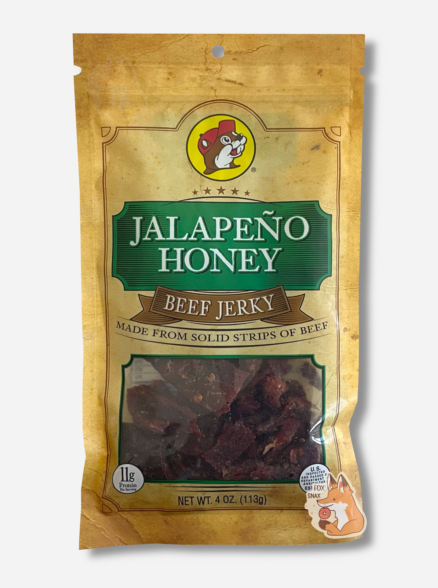 A picture of the front of a tan plastic bag of Buc-ee's Jalapeño Honey Jerky.  The logo is in green with white text.The label underneath reads 'Made From Solid Strips Of Beef'. A small window on the bottom of the bag shows the dark brown/red dried jerky inside.  The bag has a picture of Buc-ee the Beaver on it.