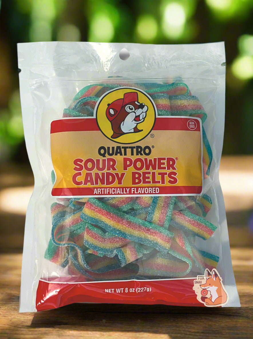 A clear square plastic bag of Buc-ee's Quattro Sour Power Candy Belts.  Inside the bag are a series of rectangular strips of candy, each about an inch and a half long.  The strips are multicolored: green, red, yellow, and blue, and are covered in white sugar.  The bag reads "QUATTRO SOUR POWER CANDY BELTS, ARTIFICIALLY FLAVORED, Net Wt 8 Oz".  The Buc-ee's logo is front and center on the bag. In upper left is a small circular logo reading "NON GMO".  The bag on a  wooden table; greenery in the background.