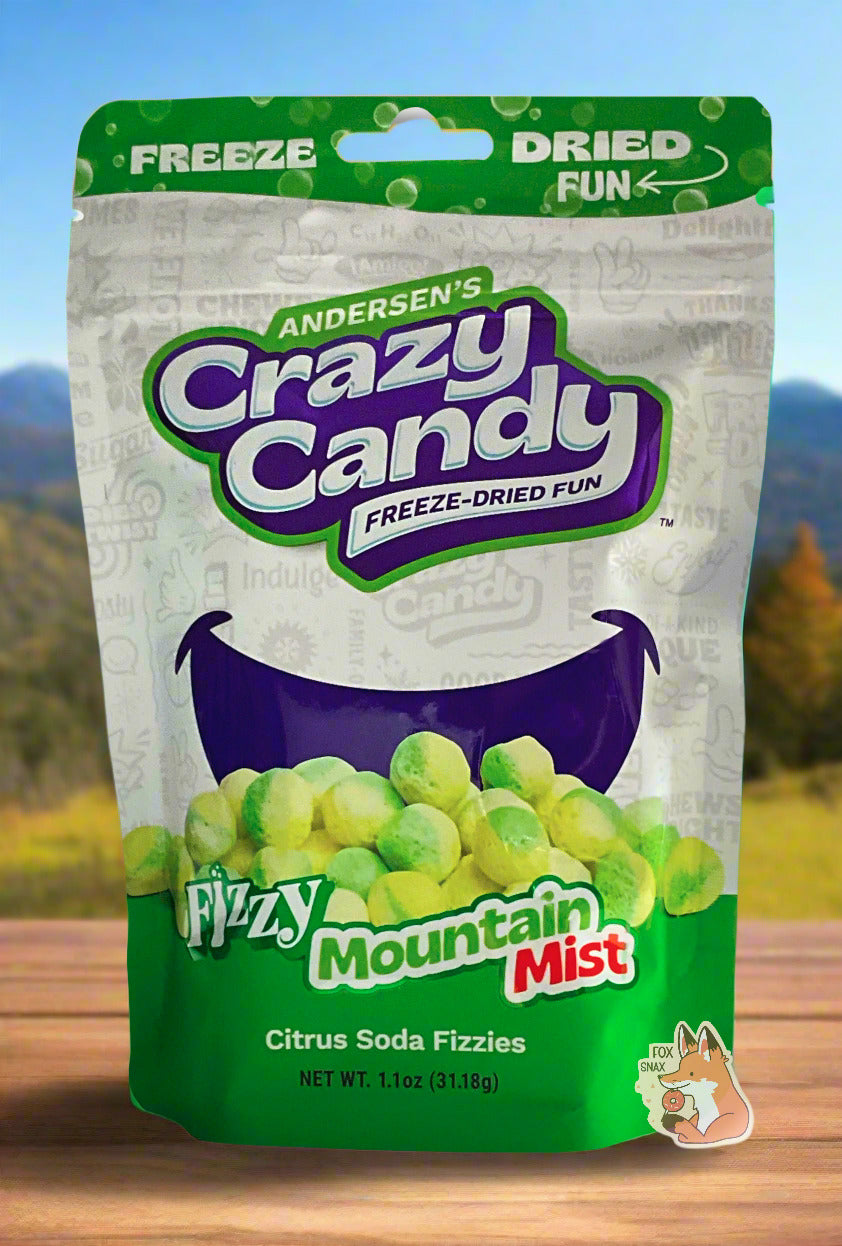 A large bag of Andersen's Fizzy Mountain Mist.  The front of the bag also says Citrus Soda Fizzies, Net Weight 1.1 oz.  It's Fun, Happy, Candy.  The top and bottom of the bag are green, and the background is a large dark blue smile against a white background.  The bag sits on a pine deck; behind the bag are distant mountains and pine trees against a pretty blue sky.