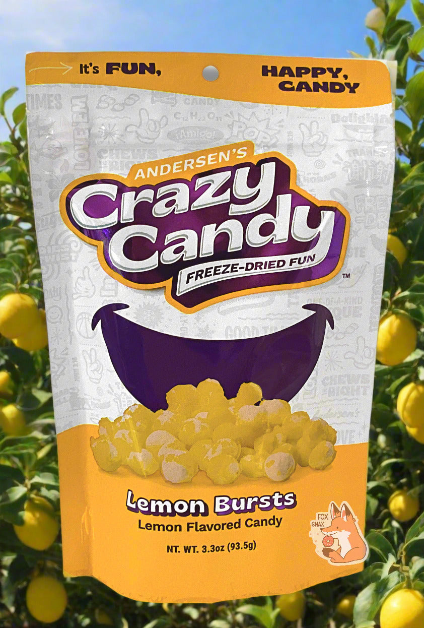 A large bag of Andersen's Lemon Bursts.  The front of the bag also says Lemon, Net Weight 3.3 oz.  It's Fun, Happy, Candy.  The top and bottom of the bag are yellow, and the background is a large purple smile against a white background.  Behind the bag is a lemon tree grove; large yellow lemons can be seen in the background next to the tree's green leaves; a blue sky frames the top of the screen.