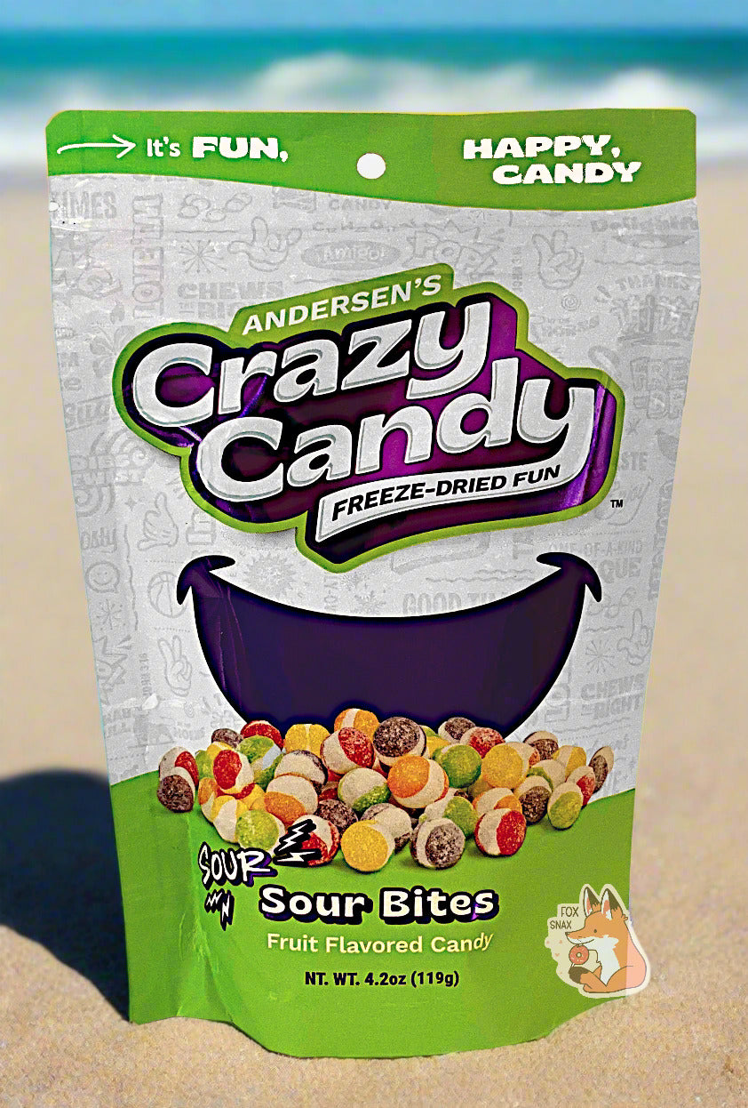 A large bag of Andersen's Sour Bites.  The front of the bag also says Sour Sour Bites, Net Weight 4.2 oz.  It's Fun, Happy, Candy.  The top and bottom of the bag are green, and the background is a large purple smile against a white background.  The bag is sitting on beach sand, with ocean waves crashing against the sand in the background against a blue sky.