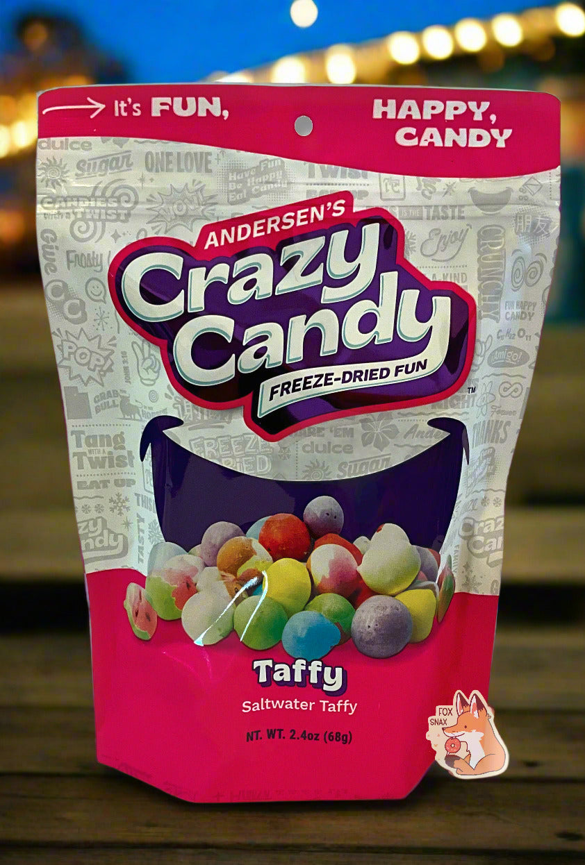 A large bag of Andersen's Taffy.  The front of the bag also says Saltwater Taffy, Net Weight 2.4 oz.  It's Fun, Happy, Candy.  The top and bottom of the bag are reddish pink, and the background is a large purple smile against a white background. The bag is sitting on a beach boardwalk at night; pretty multicolored chain lights can be seen in the background.