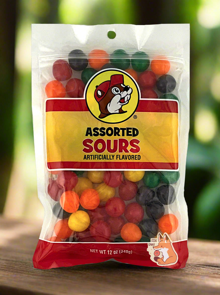 A picture of the front of a resealable bag of Buc-ee's Assorted Sours, which are sour and sweet candies.  The bag reads ASSORTED SOURS, Artificially Flavored, and has a picture of Buc-ee the Beaver on it.  The sours inside are spherical, and are multiple shades of red, orange, yellow, green, and purple.  The background is of bright green leafy foliage, with the bag on a wood table.