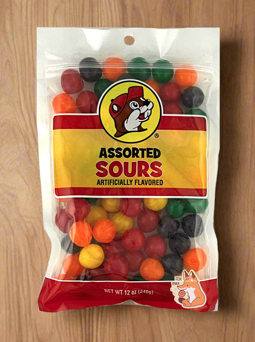 A picture of the front of a resealable bag of Buc-ee's Assorted Sours, which are sour and sweet candies.  The bag reads ASSORTED SOURS, Artificially Flavored, and has a picture of Buc-ee the Beaver on it.  The sours inside are spherical, and are multiple shades of red, orange, yellow, green, and purple.  The bag is resting on a light wood table.