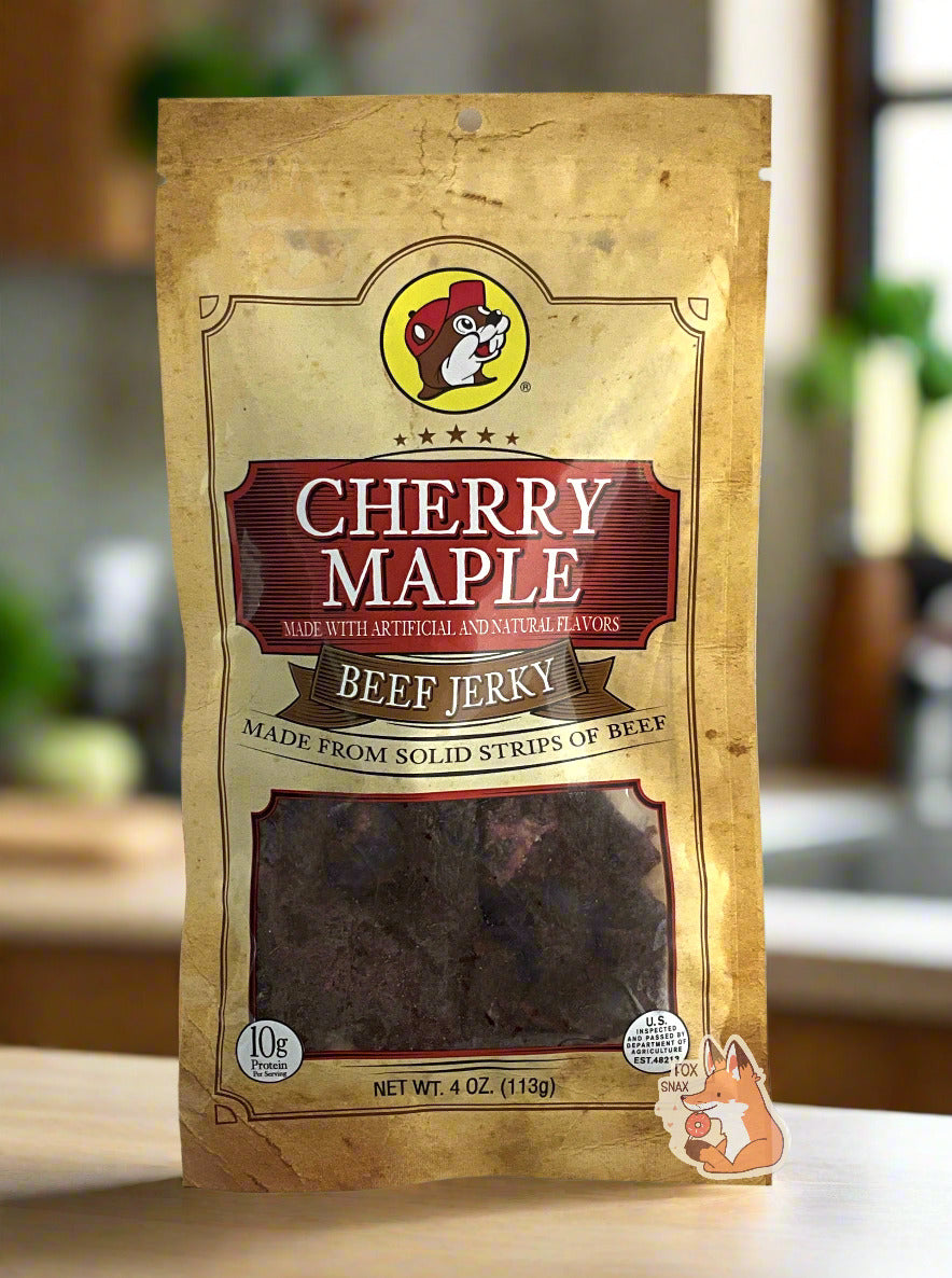 A picture of the front of a tan plastic bag of Buc-ee's Cherry Maple Beef Jerky.  The logo is in dark red with white text.The label underneath reads 'Made From Solid Strips Of Beef'.A small window on the bottom of the bag shows the dark brown/red dried jerky inside.  The bag has a picture of Buc-ee the Beaver on it.  The background is of a fancy, dark-wood paneled kitchen, with plants in the background.  The bag of jerky is resting on a light wooden table.