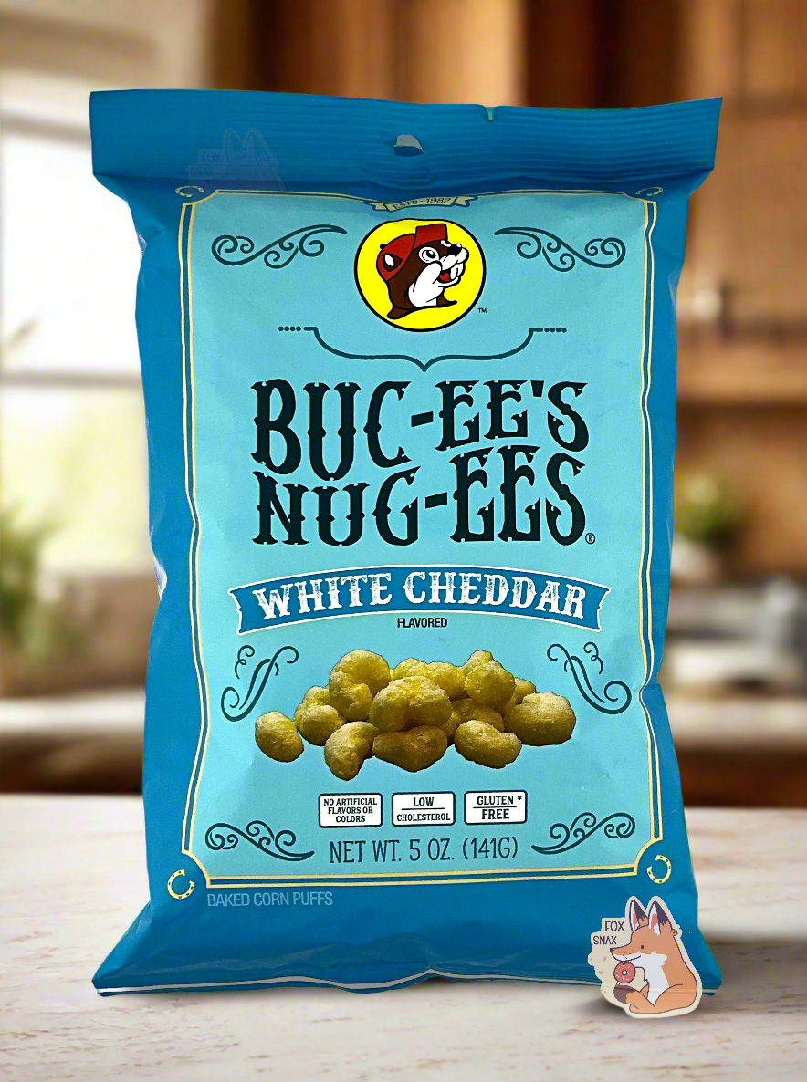 A picture of the front of a opaque, dark blue and light blue bag of Buc-ee's Nug-ees White Cheddar which are puffed corn snack with white cheddar flavor, and has a picture of Buc-ee the Beaver on it.  The bag reads White Cheddar Flavored, No Artificial Colors or Flavors, Low Cholesterol, Gluten Free, Baked Corn Puffs.  The bag is sitting on a marble countertop in a kitchen with wooden cabinets.