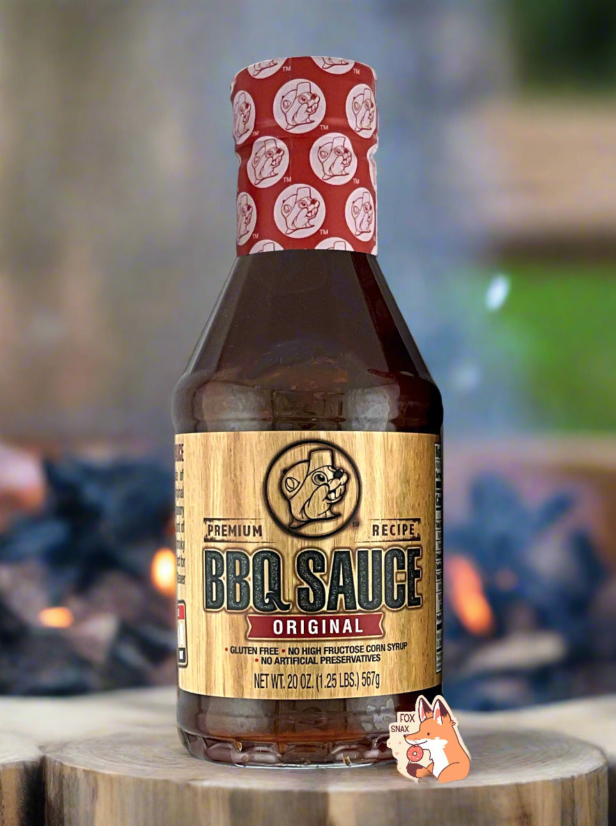 A plastic bottle of Buc-ee's Premium Recipe BBQ Sauce, Original.  The top of the bottle is sealed with a plastic wrap in red and white, dotted with Buc-ee's logos.  The BBQ label is a woodgrain, with a seared Buc-ee logo in the middle.  The rest of the bottle is clear, and dark red-brown bbq sauce can be seen.  The label reads: Gluten Free, No High Fructose Syrup, No Artificial Preservatives.  The bottle is sitting on a log; a bonfire can be seen in the background.