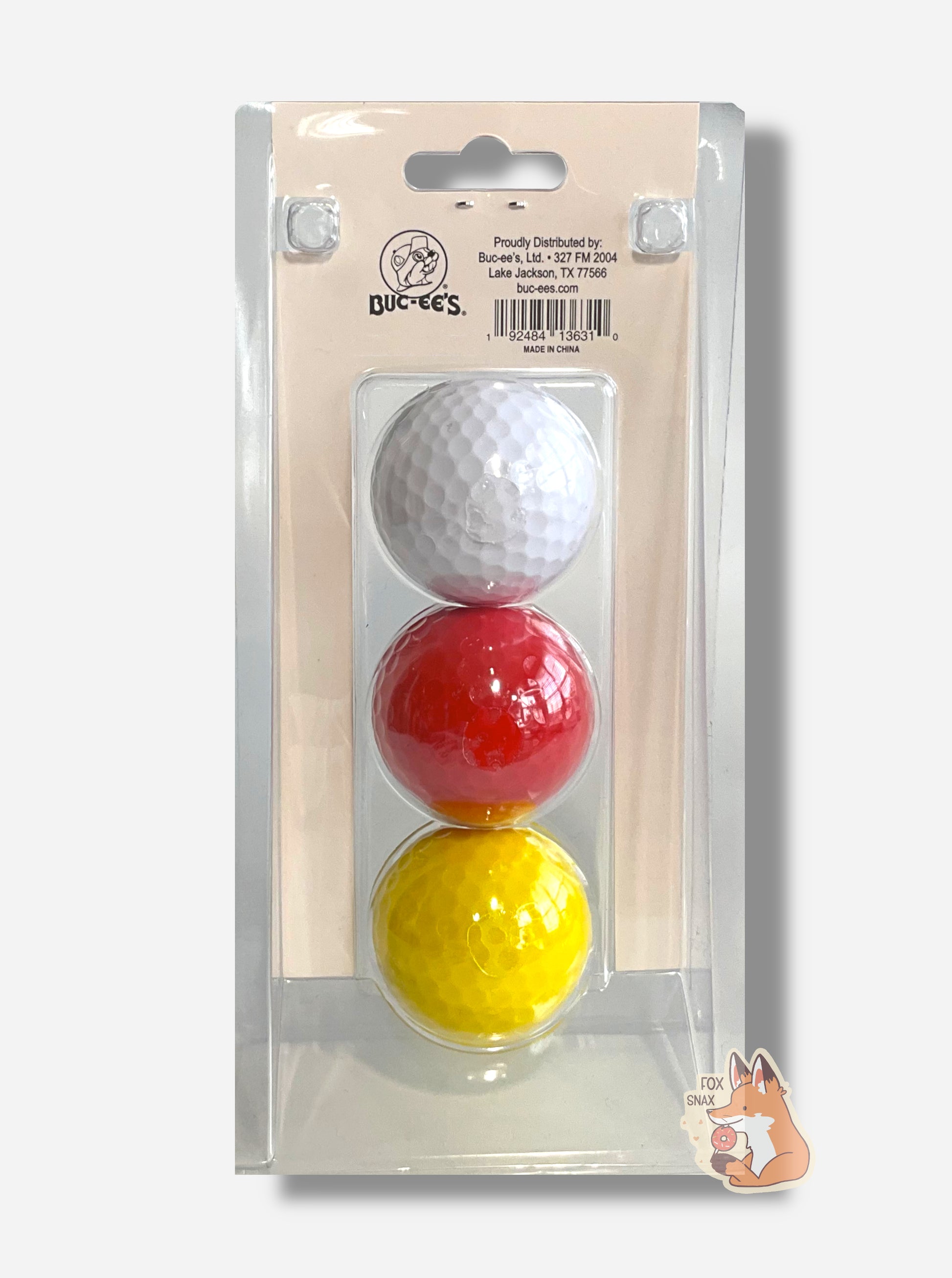A picture of the back of a rectangular, clear plastic container of three golf balls.  The golf balls are suspended in the middle of the plastic - the top one is white, middle red, and bottom yellow. This shows the back of the golf balls, and each is just a solid color on the back, with no logo.