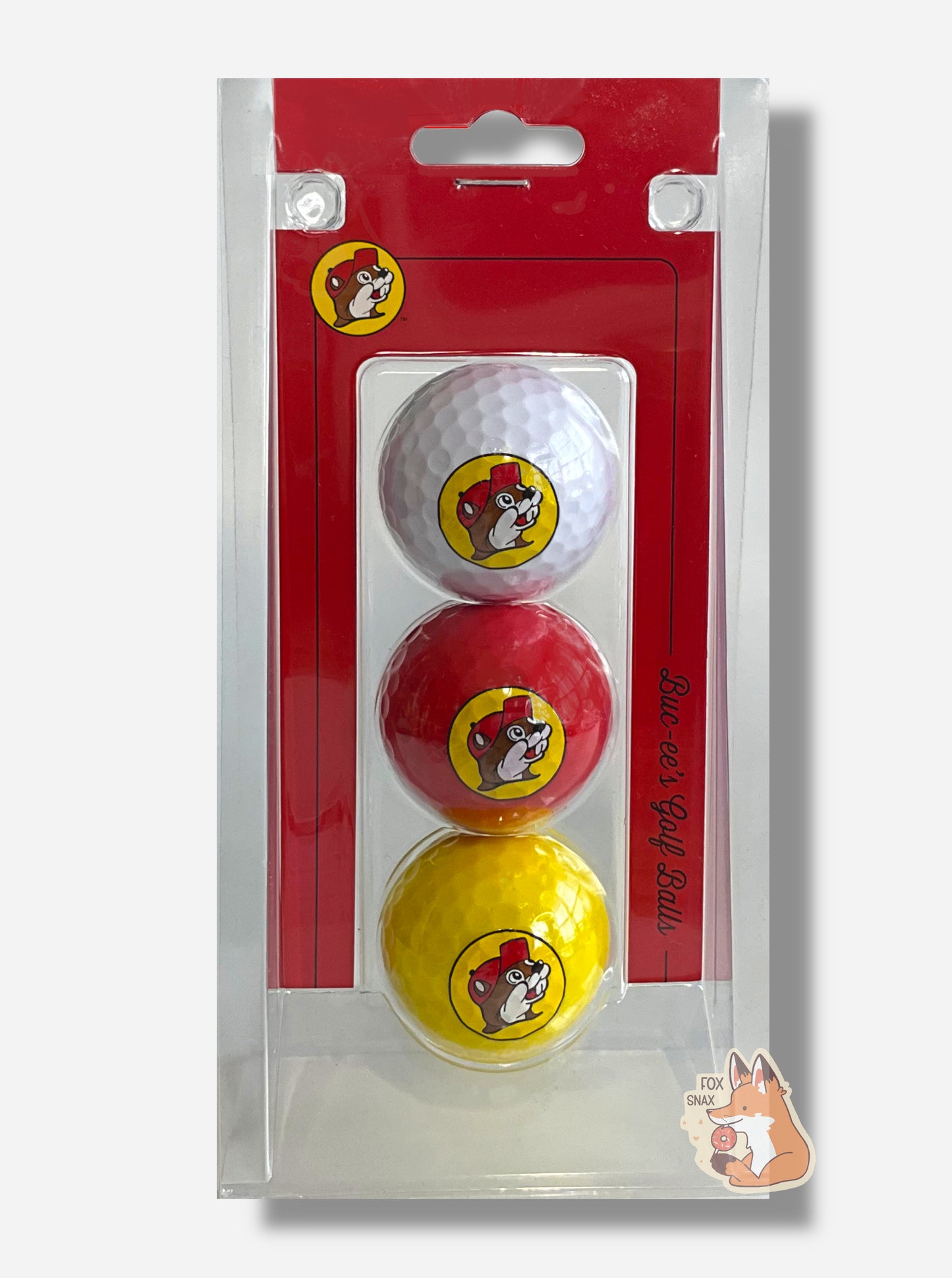 A picture of the front of a rectangular, clear plastic container of three golf balls.  The golf balls are suspended in the middle of the plastic - the top one is white, middle red, and bottom yellow.  Each golf ball has the Buc-ee's logo on it - a brown-furred beaver with a serious overbite, wearing a red ball cap.  The side of the package, in red cardboard, reads "Buc-ee's Golf Balls" in flowing black cursive script.