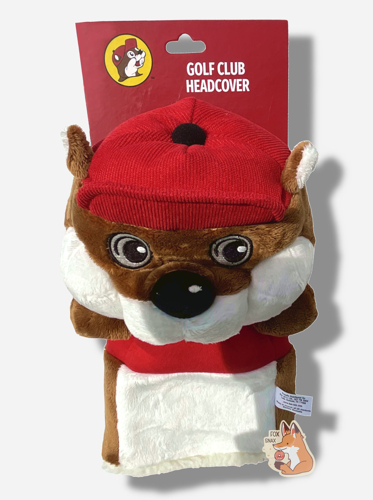 A three quarters view of Buc-ee the Beaver golf club headcover.  The head cover is of Buc-ee the beaver; a jolly cartoon beaver covered in brown and white fur, and has a big buck-tooth grin. On his head is a red ball cap, and he's wearing a red t-shirt with the Buc-ee's logo.  This golf club cover has no legs, but does have a fuzzy trim at the bottom to fit Buc-ee over a golf club.  A brown furred beaver tail can be seen hanging down from the backside.  The golf club headcover has a cardboard mounting.