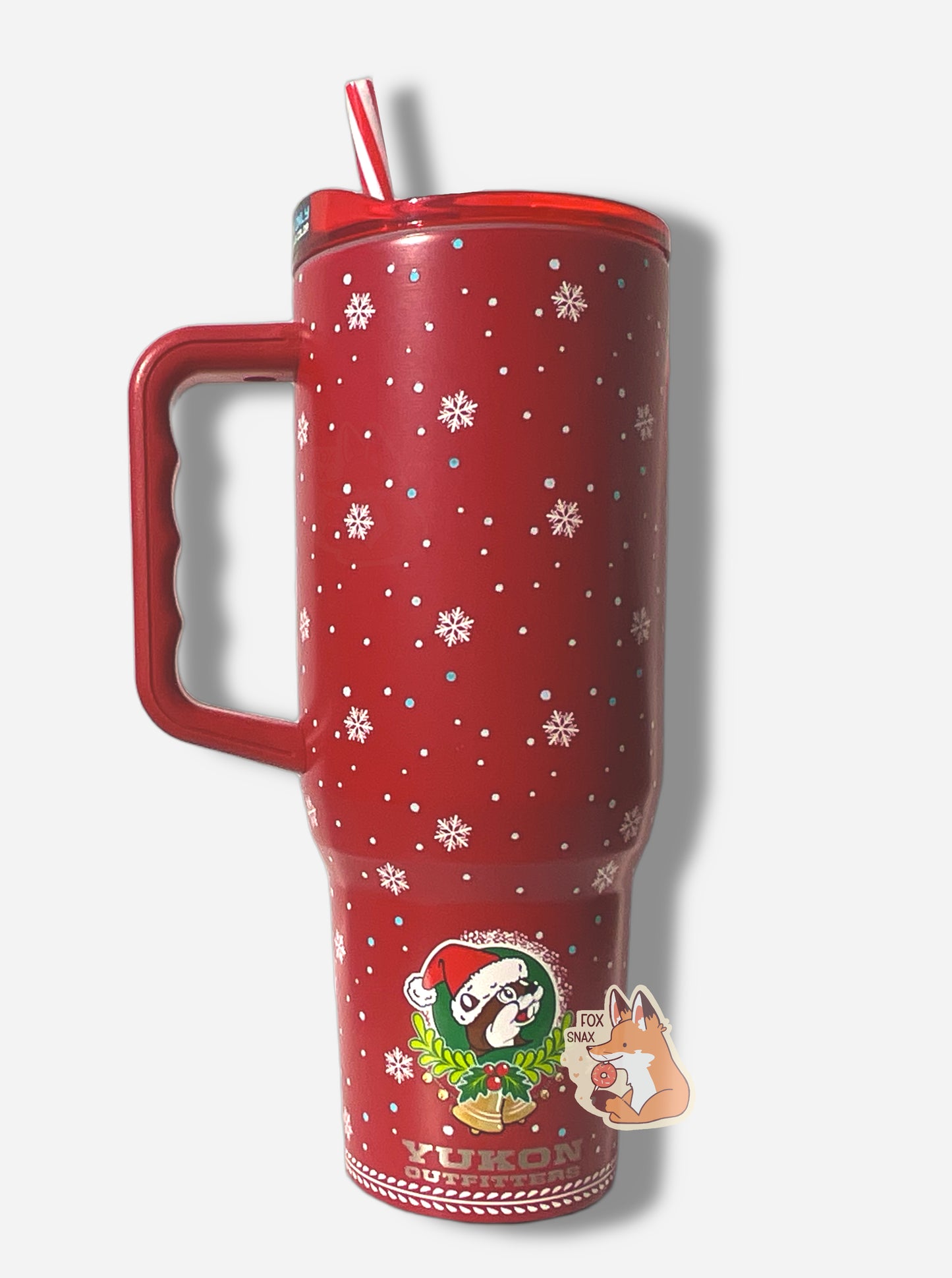 A picture of a tall red, skinny tumbler.  The tumbler is paint covering stainless steel.  The handle, top, and candy-cane colored straw are red plastic. The background around the text is darker red, with pretty white snowflakes of different sizes all around.  The bottom of the tumbler has the Buc-ee's logo; a cheerful brown-furred beaver with a significant overbite.  Buc-ee is wearing a red Christmas cap, and the bottom of the logo has a wreath with two yellow bells at the bottom.
