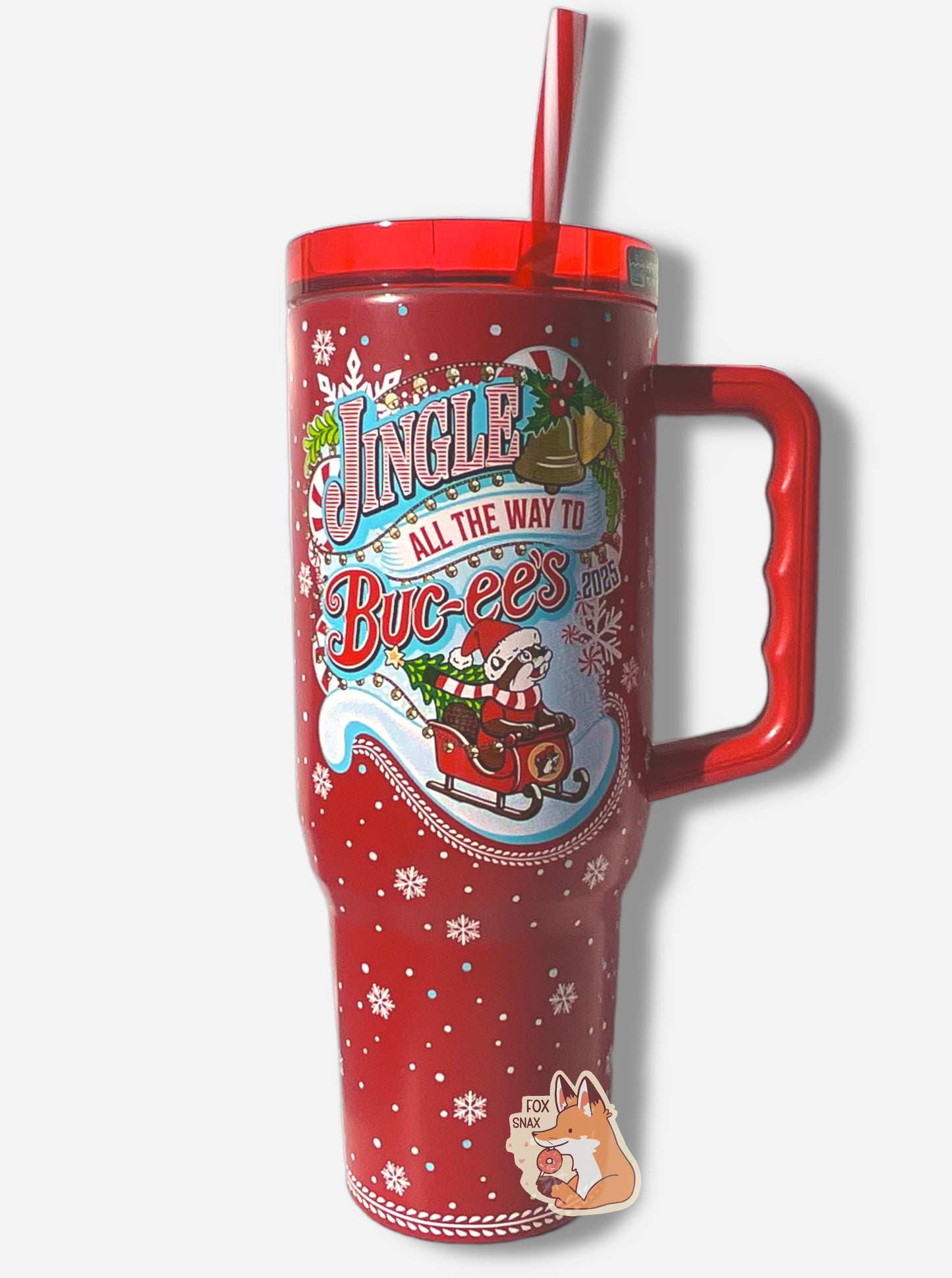 A picture of a tall red, skinny tumbler, with JINGLE ALL THE WAY TO BUC-EES written in red/white striped text. On this tumbler, Buc-ee is wearing a Christmas cap and red/white scarf, sitting in a red sleigh; the sleigh has a Christmas tree with a yellow star at the top. The tumbler is paint covering stainless steel.  The handle, top, and candy-cane colored straw are red plastic. The background around the text is darker red, with pretty white snowflakes of different sizes all around.