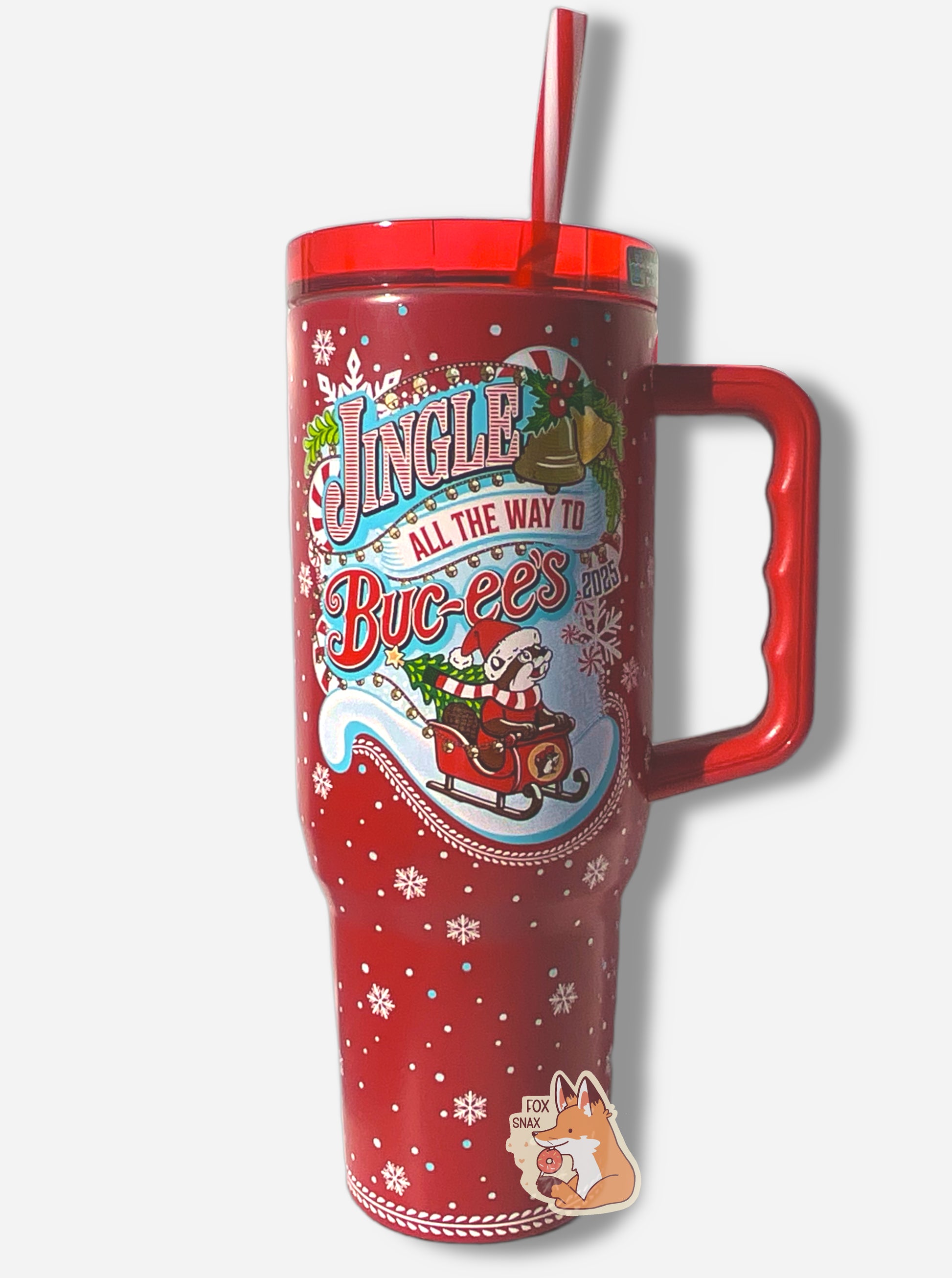 A picture of a tall red, skinny tumbler, with JINGLE ALL THE WAY TO BUC-EES written in red/white striped text. On this tumbler, Buc-ee is wearing a Christmas cap and red/white scarf, sitting in a red sleigh; the sleigh has a Christmas tree with a yellow star at the top. The tumbler is paint covering stainless steel.  The handle, top, and candy-cane colored straw are red plastic. The background around the text is darker red, with pretty white snowflakes of different sizes all around.