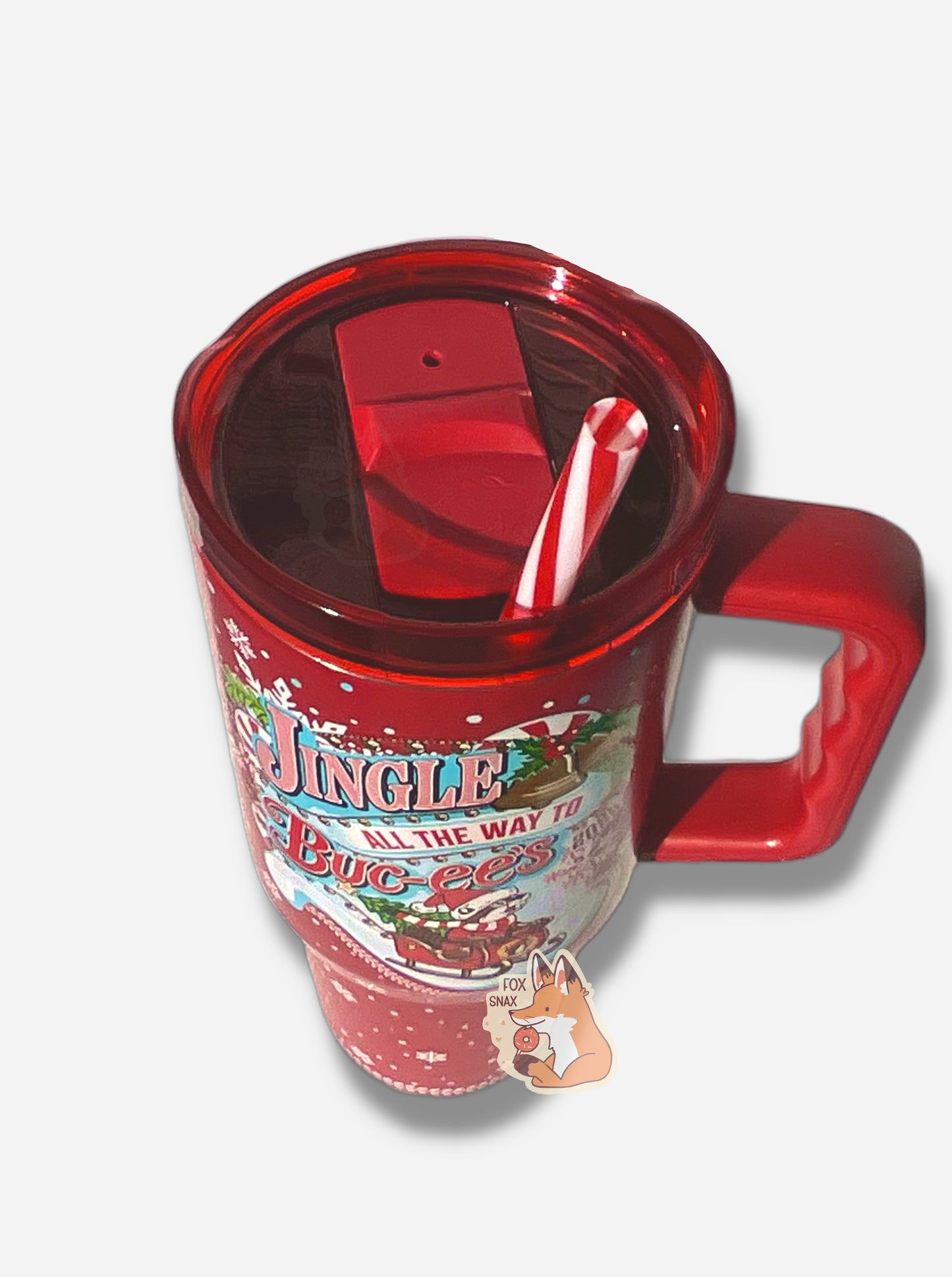 A three-quarters view of a tall red, skinny tumbler, with JINGLE ALL THE WAY TO BUC-EES written in red/white striped text. On this tumbler, Buc-ee is wearing a Christmas cap and red/white scarf, sitting in a red sleigh; the sleigh has a Christmas tree with a yellow star at the top. The tumbler is paint covering stainless steel.  The handle, top, and candy-cane colored straw are red plastic. The background around the text is darker red, with pretty white snowflakes of different sizes all around.
