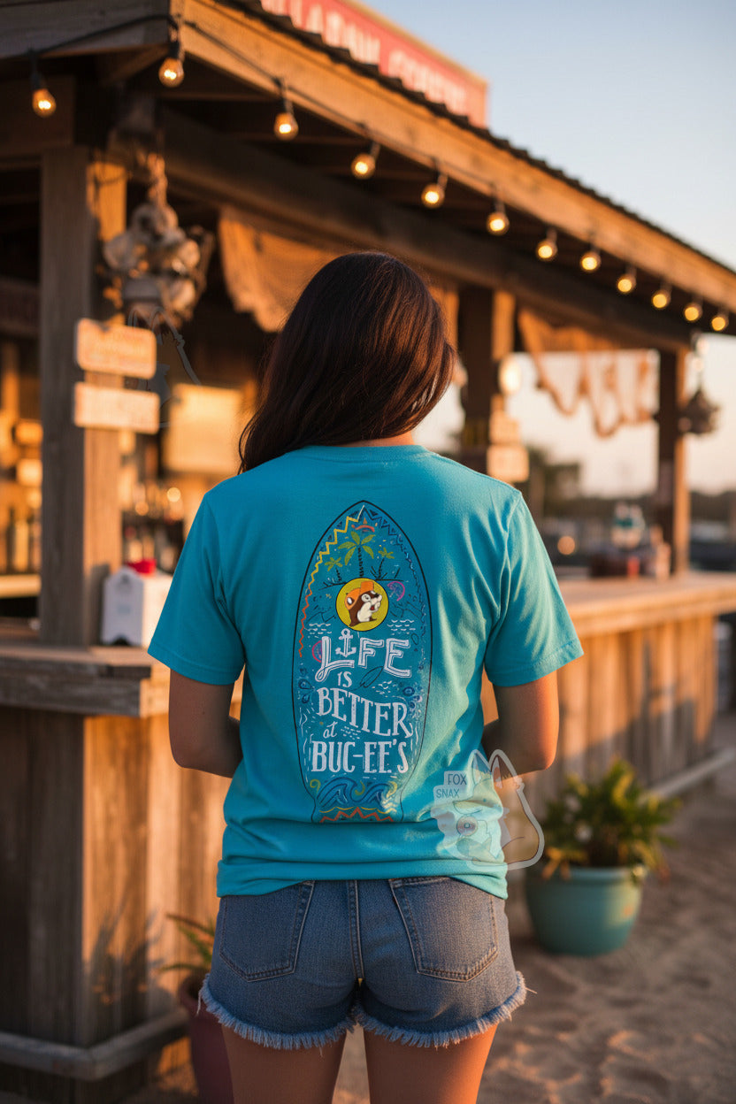 A photo of a woman with long brown hair wearing a cyan t-shirt.  The back of the shirt has a surfboard logo in black, with palm tree icons in green and black at the top, then the Buc-ee's logo, wearing an orange ballcap.  The woman is also wearing jean shorts, and is standing by a beach bar, on a beach at sunset, with her back towards the viewer.  The bar is wood-framed and is covered in lights at the top.