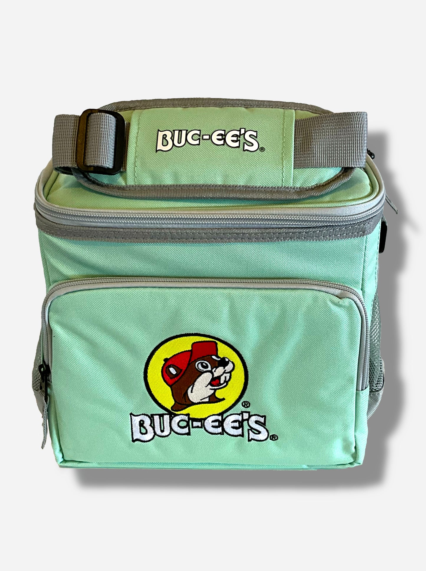 A picture of a three-quarters view of the front of a lunch box cooler.  The outer surface is made of mint colored polyester, and has a large mint colored strap on it that reads BUC-EE'S in white text.  The front of the lunch box has a zip pouch, with the Buc-ee's logo on it: a grinning beaver wearing a red hat against the background of a yellow circle.  The sides of the tote has mesh pouches, the top sealed by a zipper.