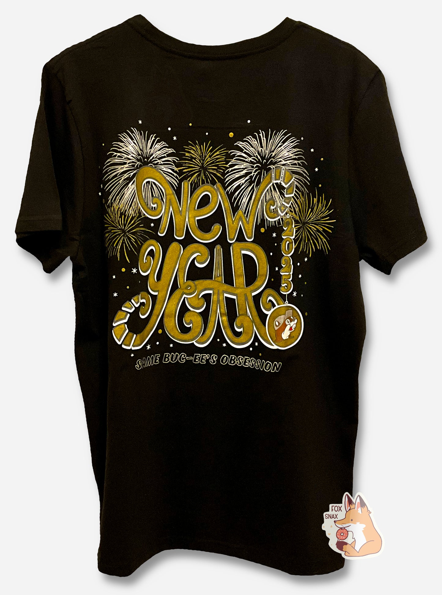 A picture of the back of a black t-shirt.  The center of the t-shirt reads in a curly gold font, NEW YEAR 2025, then underneath of it, SAME BUC-EE'S OBSESSION.  The bottom right of the shirt has the Buc-ee's logo, in gold, where red would normally be.  The background behind the text is a series of gold and silver exploding fireworks, dots, and stars.