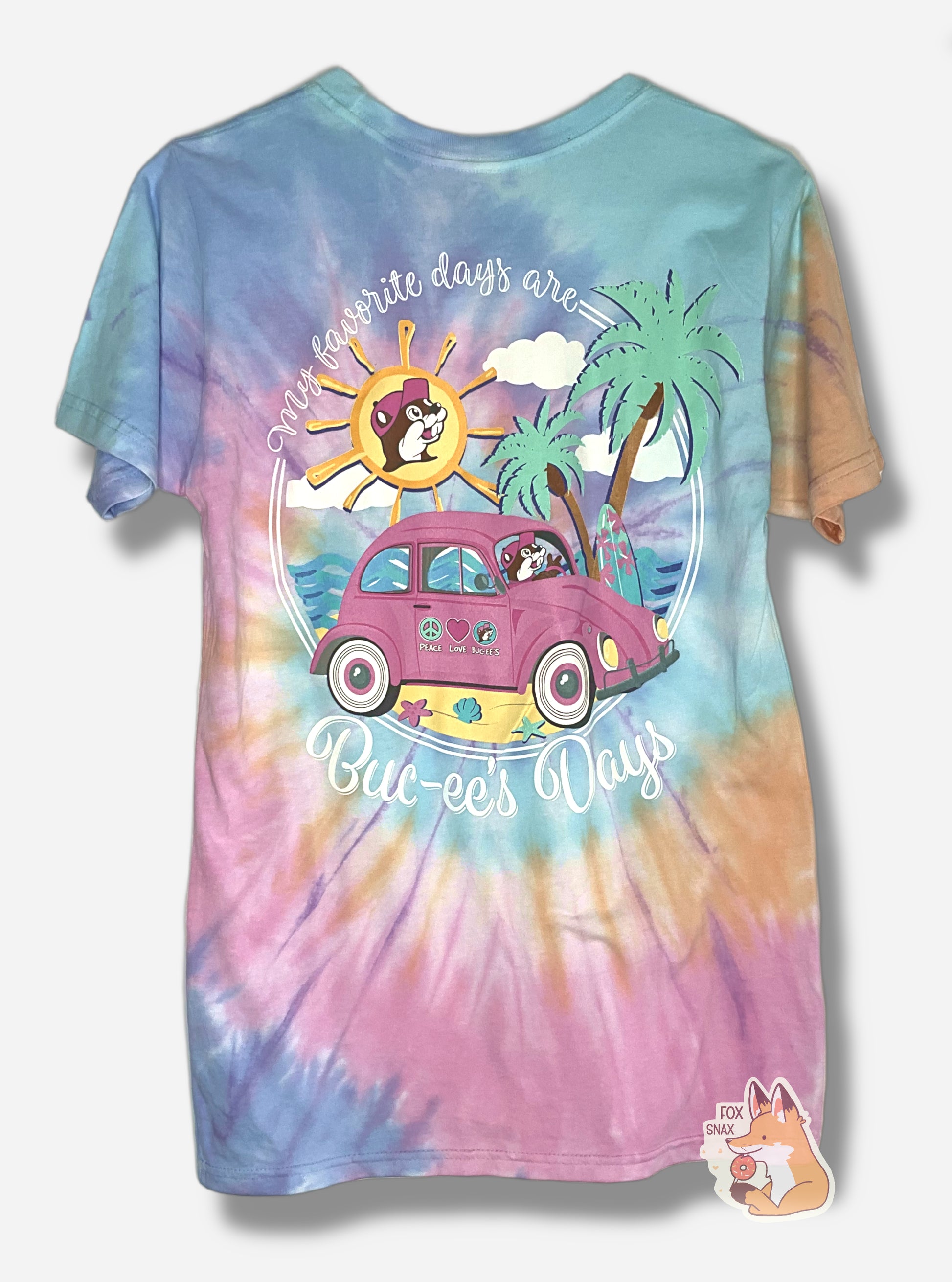 A picture of the back of a multicolor tie-dye t-shirt.  The shirt has a spiral of color; orange, blue, light blue,yellow, pink. On the back is white cursive text that reads, on the top, MY FAVORITE DAYS ARE, and on the bottom, BUC-EE'S DAYS. The center of the shirt has a pink VW Bug with white-wall tires, driving on a shell-studded beach; blue water and two palm trees can be seen in the background.  Buc-ee is driving the vehicle, wearing a dark pink ball cap.  The sky is multicolored and has a surreal sun.