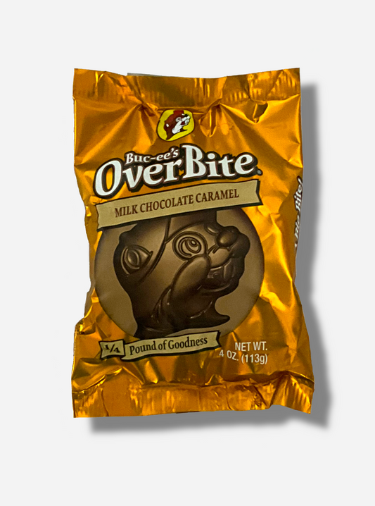 A picture of a package of a milk chocolate caramel Buc-ee's OverBite.  The package is metallic and orange/tan, and features a large picture of the candy, which is shaped like the Buc-ee's beaver mascot logo.  The package reads BUC-EE's OVERBITE, then MILK CHOCOLATE CARAMEL, then 1/4 POUND OF GOODNESS.