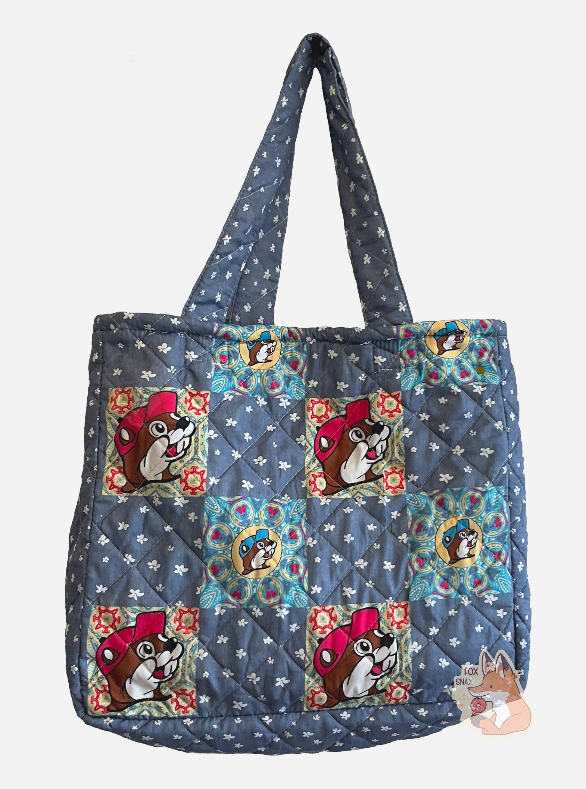 A picture of the front of a Buc-ee's Patchwork Tote.  The tote is a large, rectangular. flat bag.  The front and back of the bag are blue cloth, peppered with very small white flowers.  On four positions are an imprint of Buc-ee The Beaver's face with a pink cap, against a square colorful background.  Four other positions have smaller version of Buc-ee with light blue caps against a blue and yellow square background.  Two cloth straps are sewn to the top of the bag to make it easy to carry.