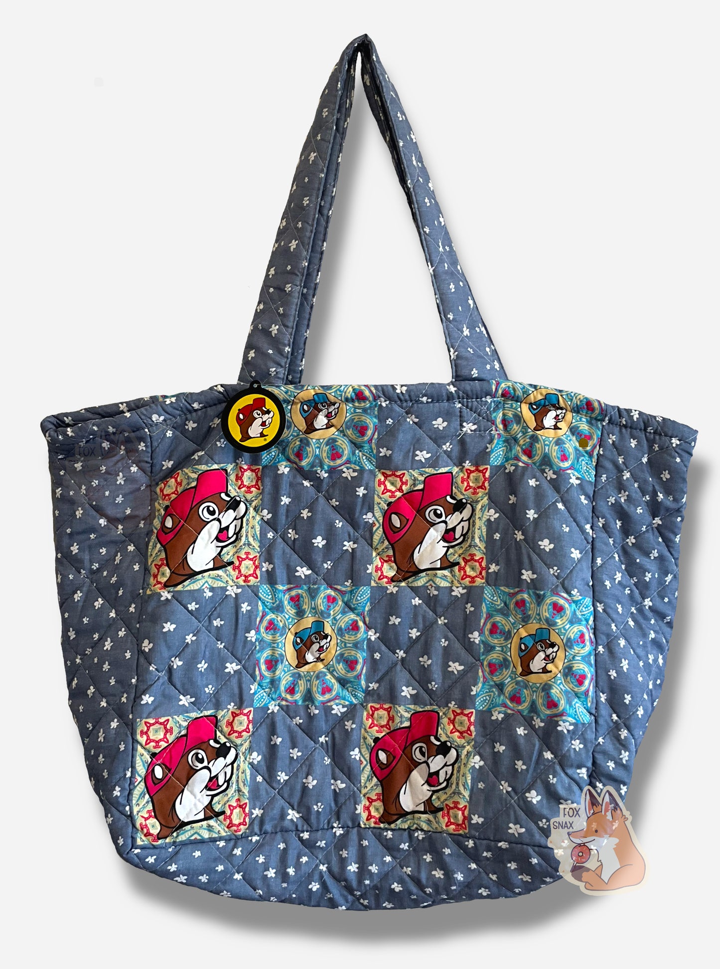 A picture of the front of a Buc-ee's Patchwork Tote.  The tote is a large, rectangular. flat bag.  The front and back of the bag are blue cloth, peppered with very small white flowers.  On four positions are an imprint of Buc-ee The Beaver's face with a pink cap, against a square colorful background.  Four other positions have smaller version of Buc-ee with light blue caps against a blue and yellow square background.  Two cloth straps are sewn to the top of the bag to make it easy to carry.