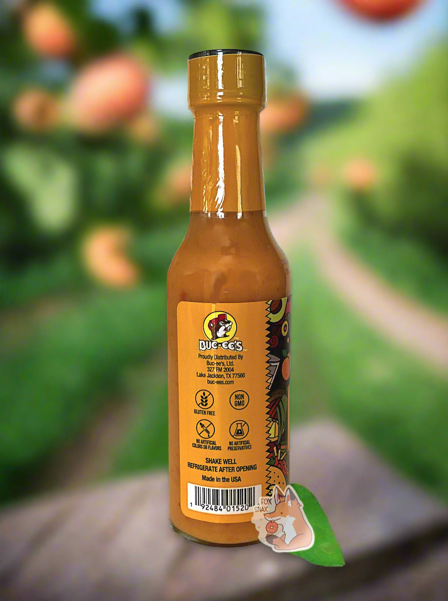A picture of a small, tall glass bottle of hot sauce. The top of the bottle is sealed with a plastic top.  The back reads GLUTEN FREE, NON GMO, NO ARTIFICIAL COLORS OR FLAVORS, NO ARTIFICIAL PRESERVATIVES, then SHAKE WELL and REFRIGERATE AFTER OPENING.  Made in the USA.  The sauce inside is bright orange.  The background if of a peach grove, with fresh ripe peaches available for picking off a branch.