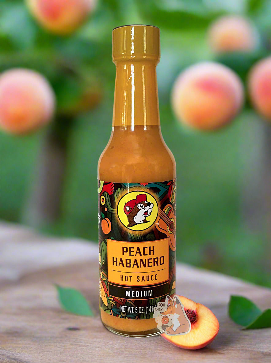 A picture of a small, tall glass bottle of hot sauce.  The top of the bottle is sealed with a orange-yellow plastic cover on a black cap.  The front label of the bottle reads PEACH HABANERO HOT SAUCE, FIERY HOT.  The Buc-ee's logo can be seen on the front; the background is a fairly busy abstract artwork featuring a guitar, a peach, and a taco.  The sauce inside is bright orange / yellow.  The background if of a peach grove, with fresh ripe peaches available for picking off a branch.