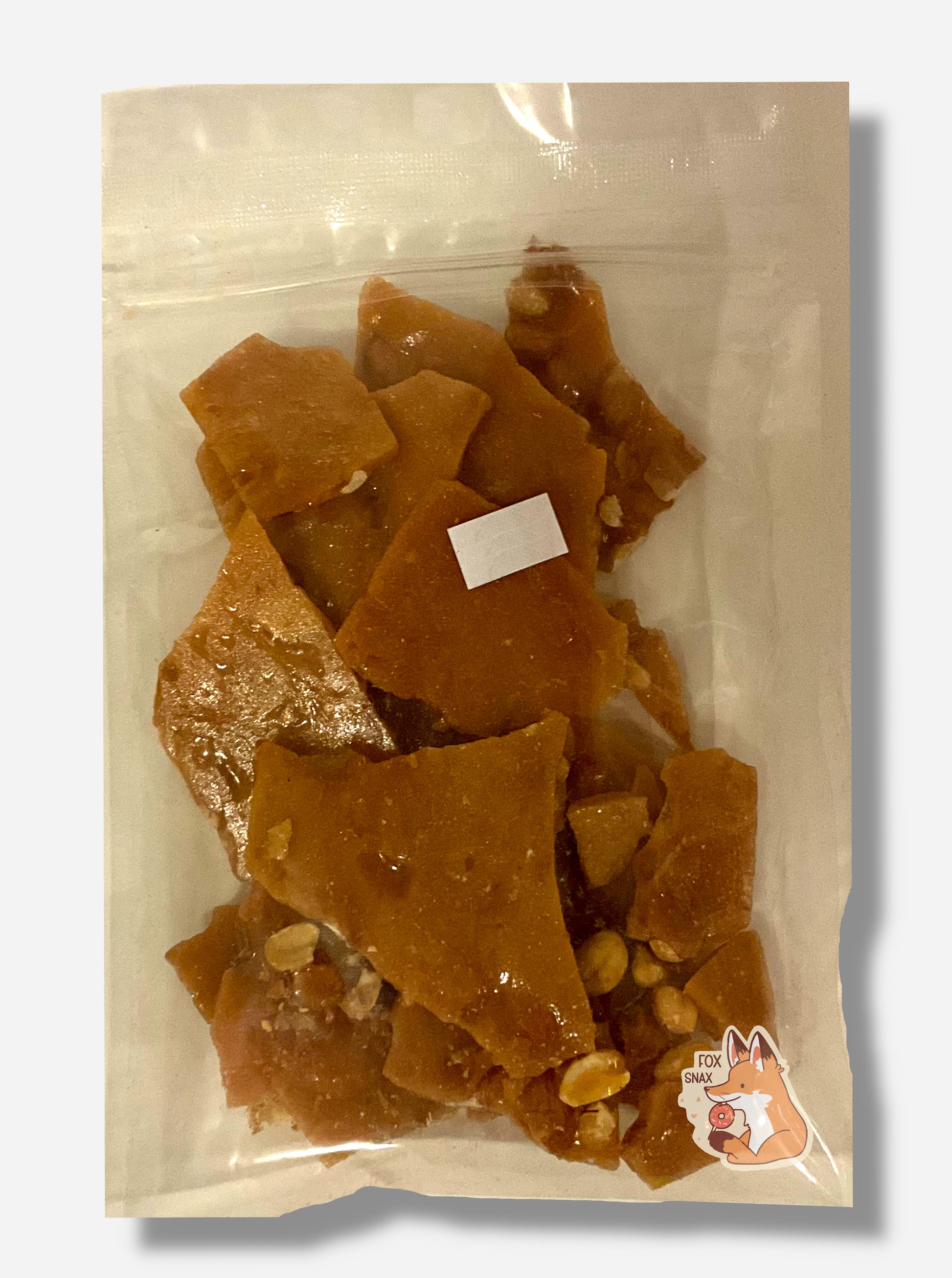 A picture of the back of a small clear plastic bag of Buc-ee's Handmade Peanut Brittle.  Inside the plastic bag are brownish irregular shapes, the brittle, intermittently dotted with pieces of peanuts.