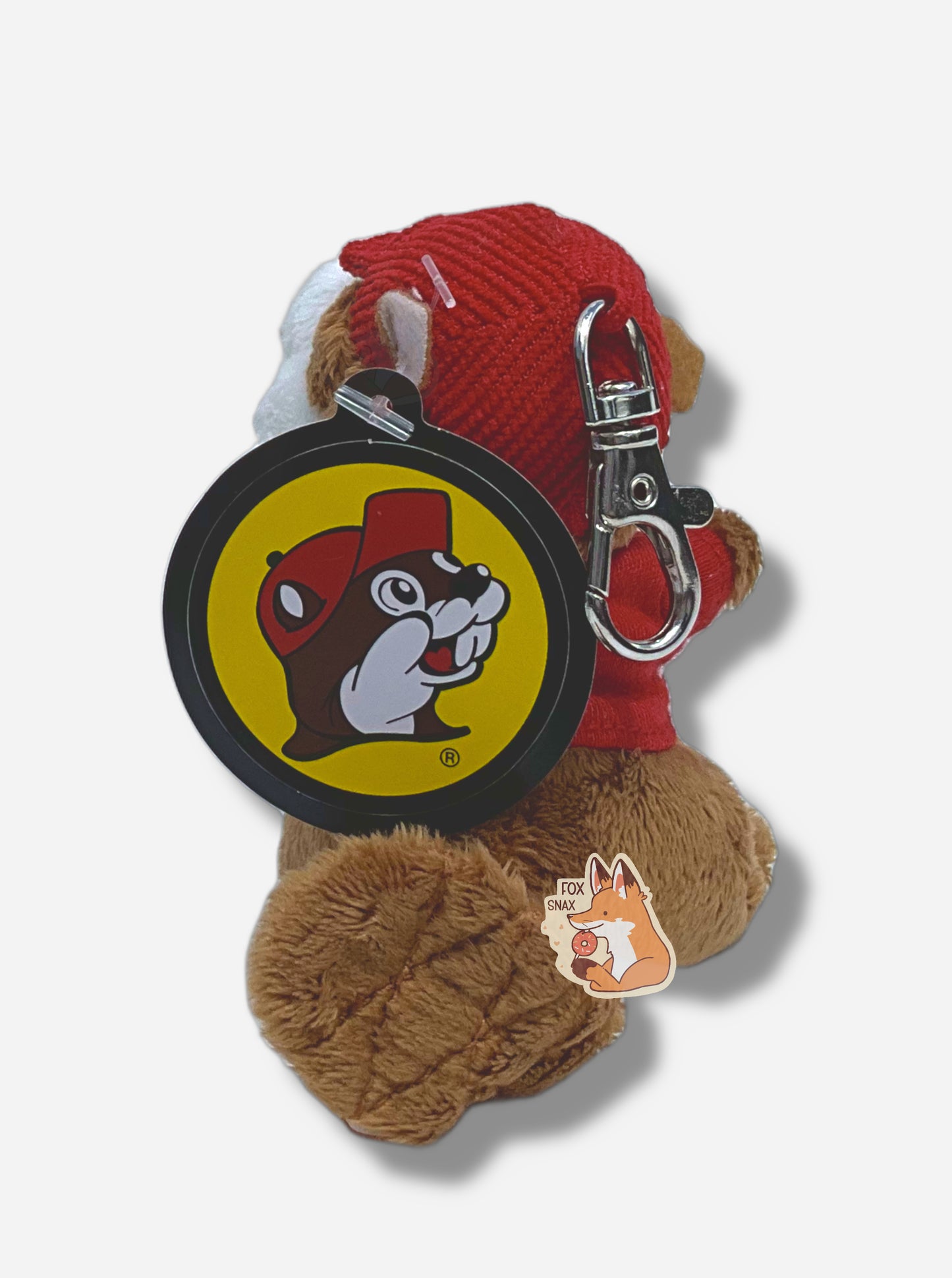 A picture of the back of a small fuzzy Buc-ee the Beaver keychain.  The keychain is made of fuzzy fur-like material, and is of Buc-ee the Beaver:  a brown-furred beaver with a serious overbite and fat beaver tail, wearing a red Buc-ee's t-shirt and a red ball cap.  The back of the hat has a small metal clasp to hook the keychain to the rest of one's keys.