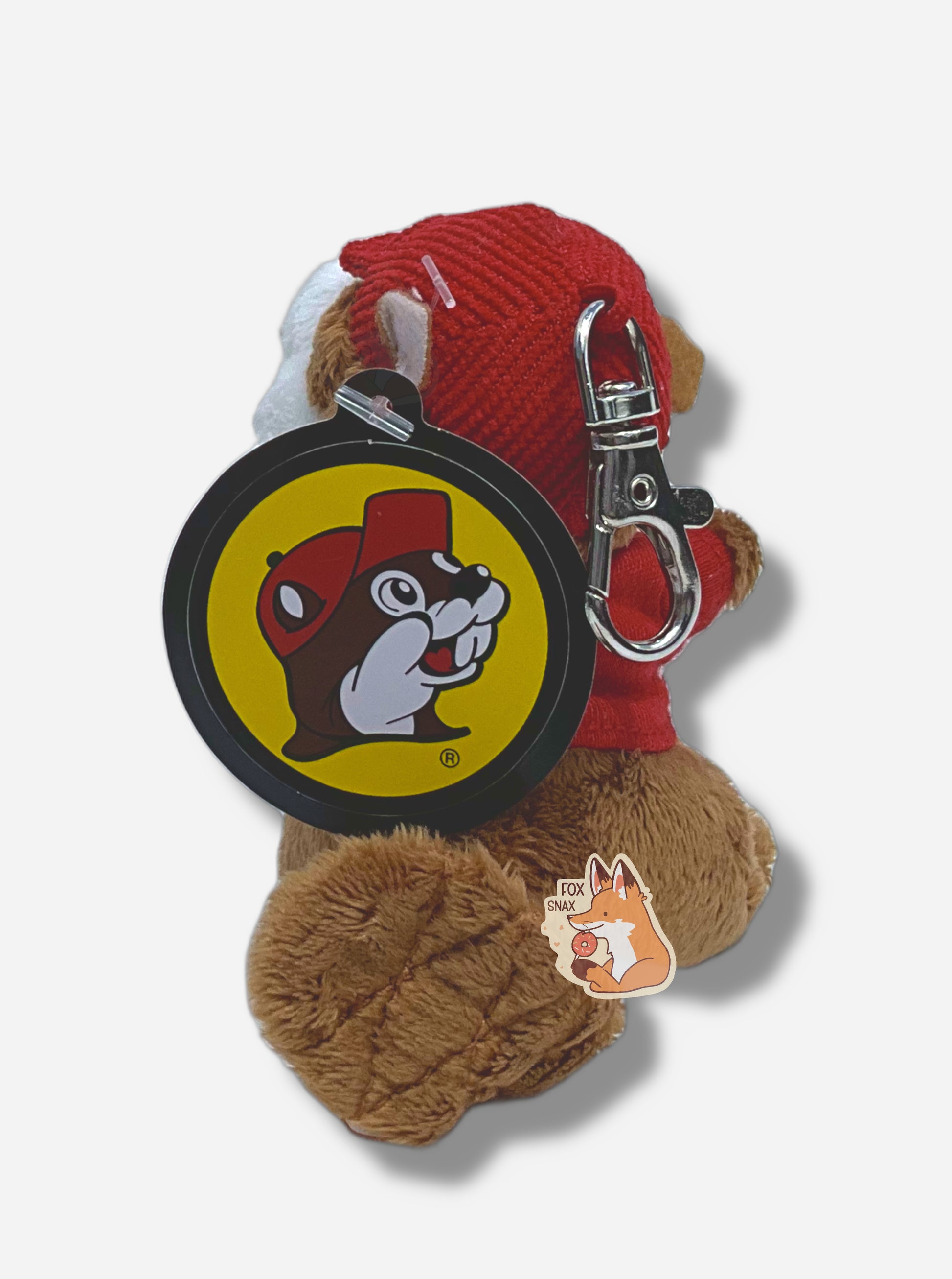 A picture of the back of a small fuzzy Buc-ee the Beaver keychain.  The keychain is made of fuzzy fur-like material, and is of Buc-ee the Beaver:  a brown-furred beaver with a serious overbite and fat beaver tail, wearing a red Buc-ee's t-shirt and a red ball cap.  The back of the hat has a small metal clasp to hook the keychain to the rest of one's keys.