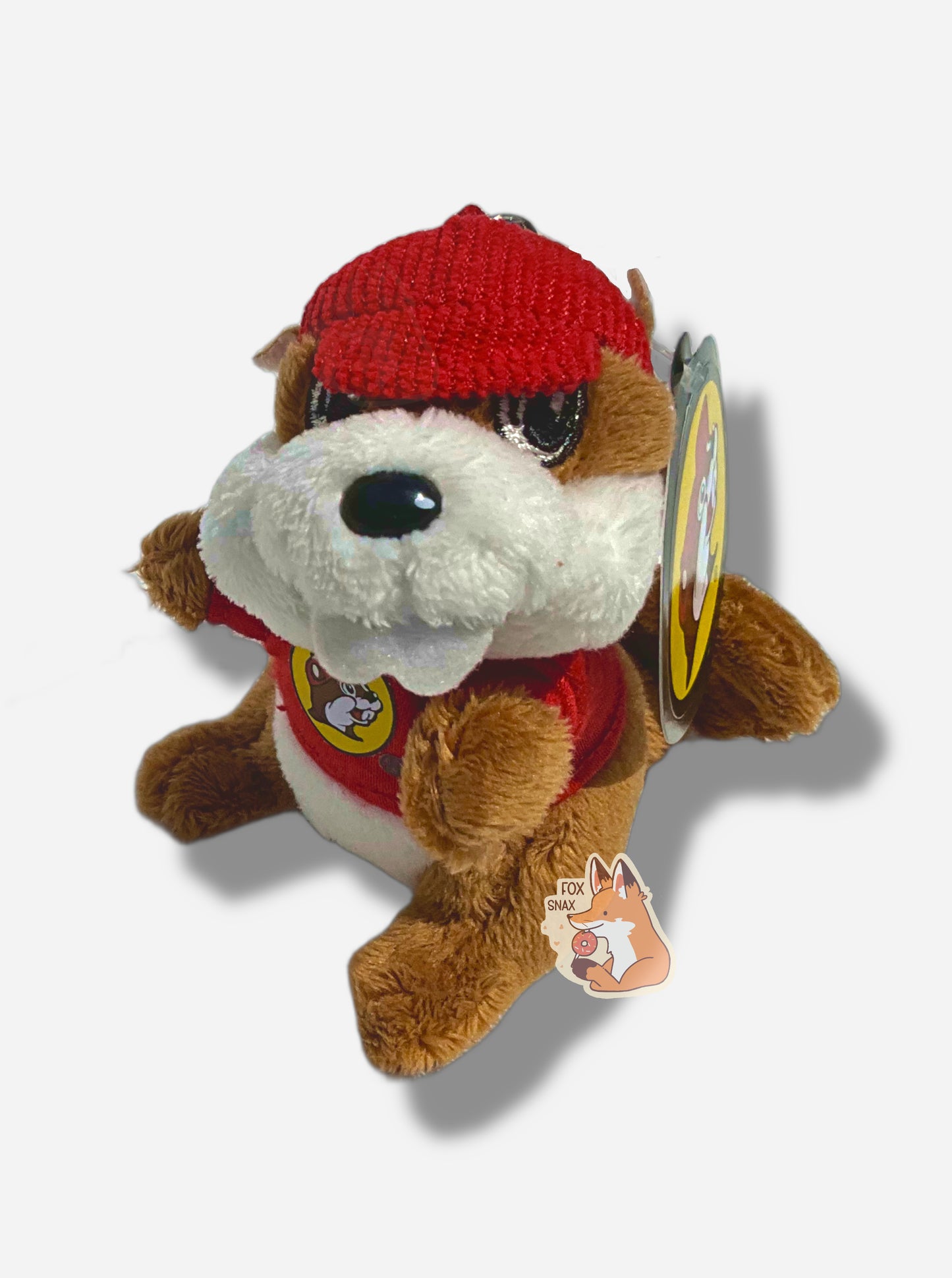 A picture of a small fuzzy Buc-ee the Beaver keychain.  The keychain is made of fuzzy fur-like material, and is of Buc-ee the Beaver:  a brown-furred beaver with a serious overbite and fat beaver tail, wearing a red Buc-ee's t-shirt and a red ball cap.  The back of the hat has a small metal clasp to hook the keychain to the rest of one's keys.