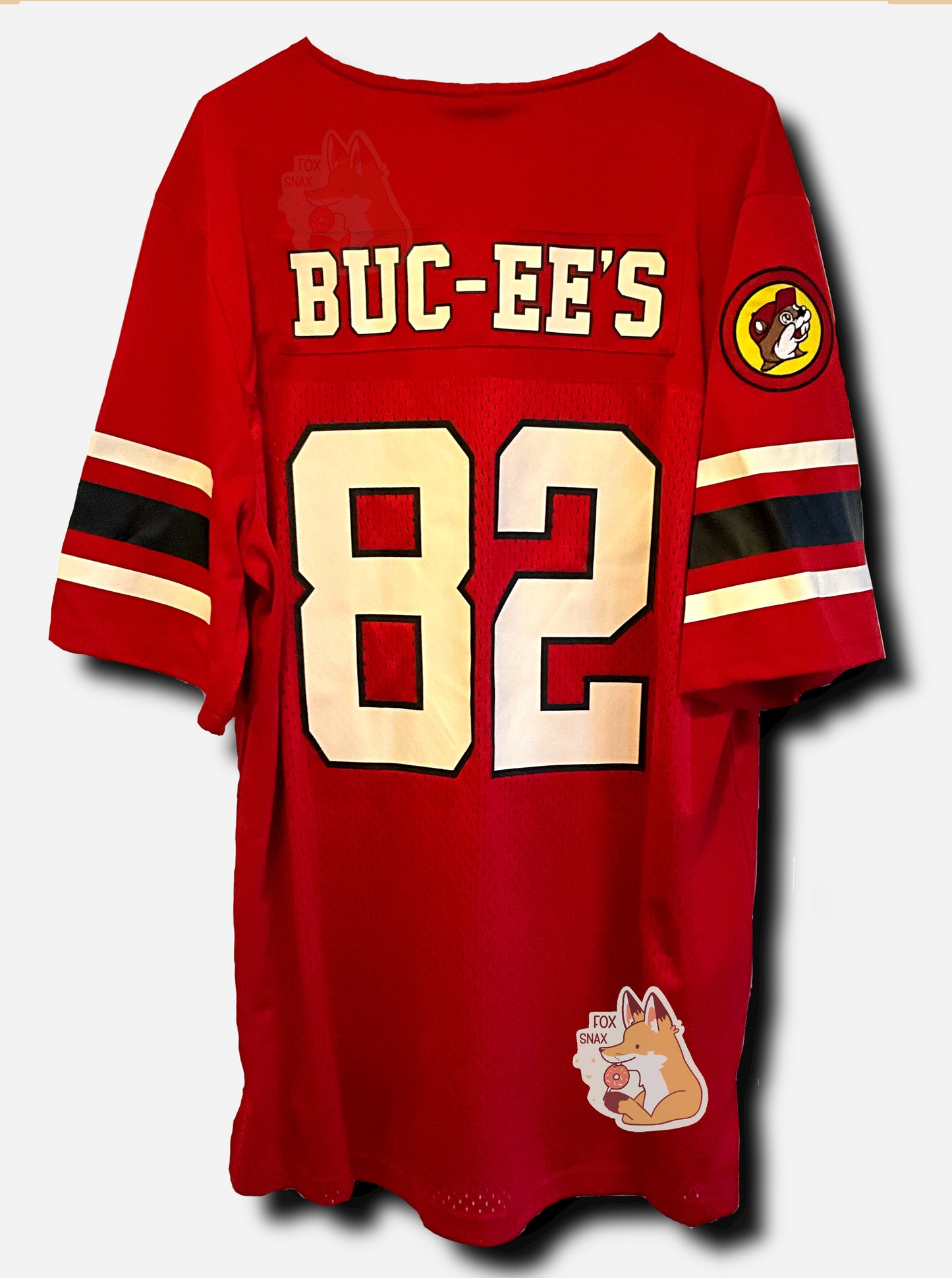 A picture of a the back of red football jersey.  On the back of the jersey, it reads BUC-EE'S, then underneath that, a large 82 for the number.  The arms have two white and one black stripe.  The right arm has a Buc-ee's patch with Buc-ee in a yellow circle, wearing a white hat.  