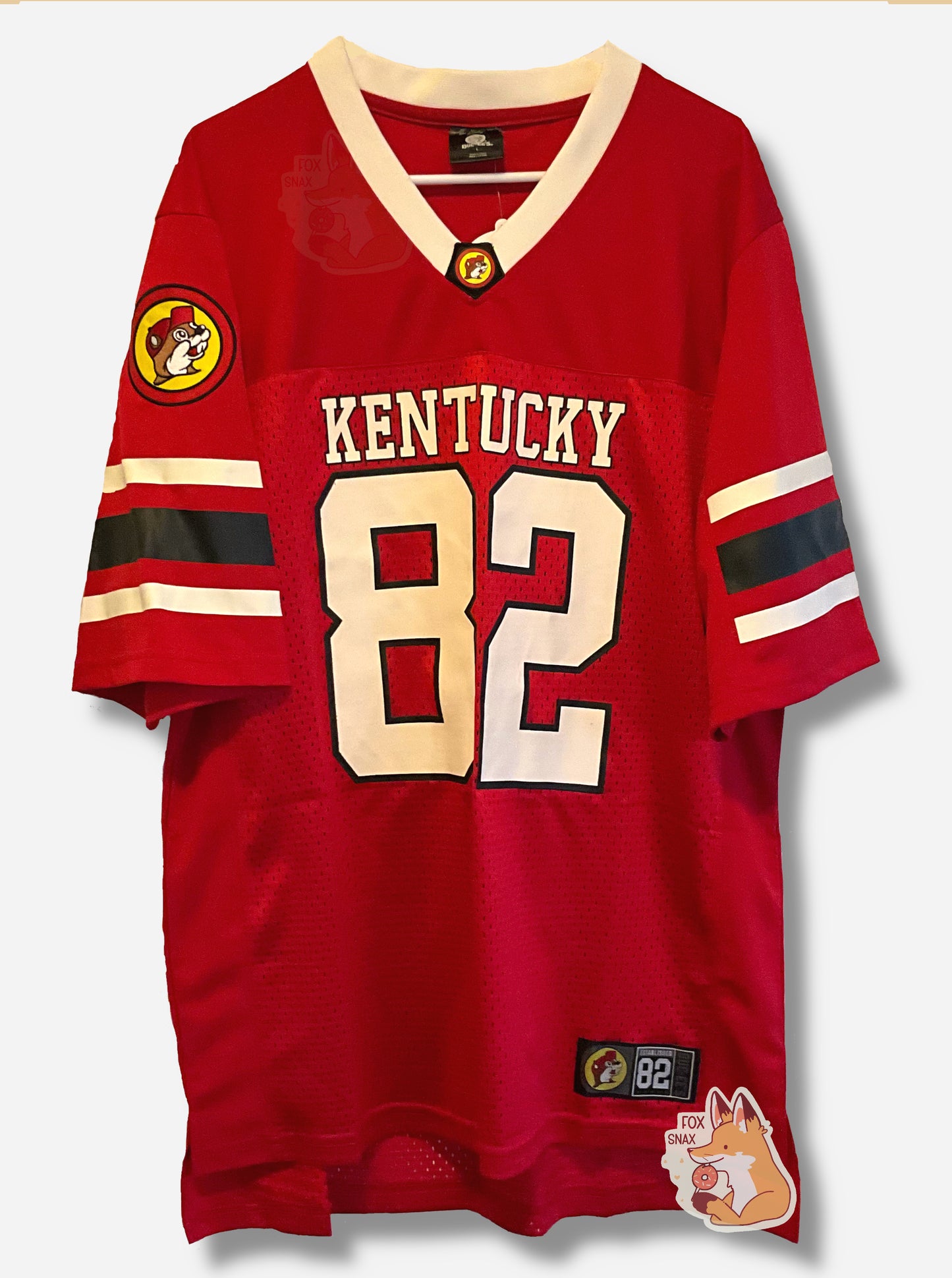 A picture of a the front of a red football jersey.  On the chest of the jersey, it reads KENTUCKY, then underneath that, a large 82 for the number.  The arms have two white and one black stripe.  The right arm has a Buc-ee's patch with Buc-ee in a yellow circle, wearing a white hat.  The neckline is a V-shape, and in white. A small rectangular patch on the bottom shirt reads BUC-EES ESTABLISHED 82