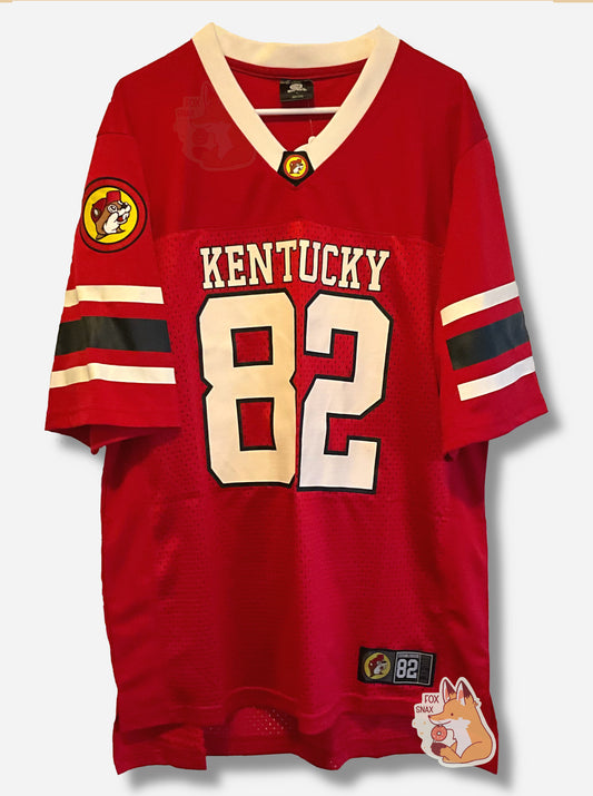 A picture of a the front of a red football jersey.  On the chest of the jersey, it reads KENTUCKY, then underneath that, a large 82 for the number.  The arms have two white and one black stripe.  The right arm has a Buc-ee's patch with Buc-ee in a yellow circle, wearing a white hat.  The neckline is a V-shape, and in white. A small rectangular patch on the bottom shirt reads BUC-EES ESTABLISHED 82