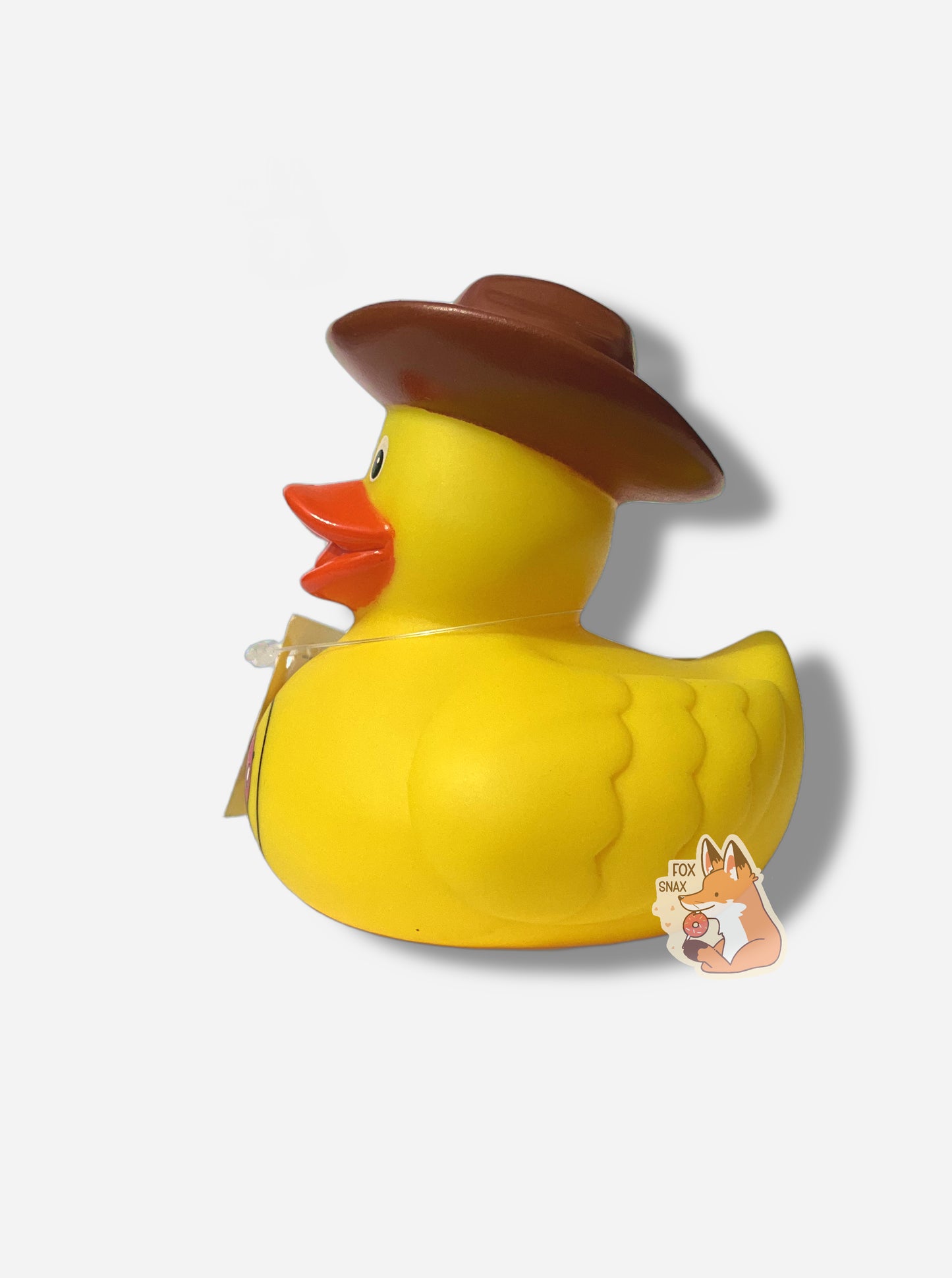 A picture of the side of a yellow rubber duck.  The duck has an orange beak, and is made of plastic.  It has a orange beak, imprints of feathers near its tail, and is wearing a brown cowboy hat on its head.