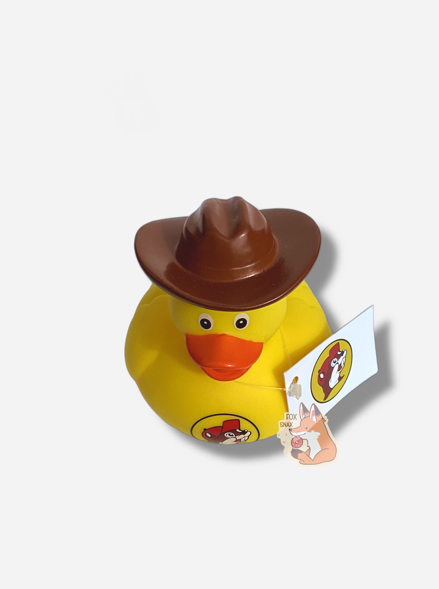 A three quarters view of a yellow rubber duck wearing a large brown cowboy hat. The duck has an orange beak, and is made of plastic.  It is wearing a dark brown hat on its head, and a red t-shirt around its body.  The front of the t-shirt has the Buc-ee's logo: a cheerful, brown furred beaver with a significant overbite, which is wearing a red ball cap.  A small white rectangular price tag is around its neck.
