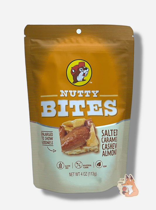 A picture of a white and purple bag of Buc-ee's Salted Caramel Cashew Almond Nutty Bites. The Buc-ee's logo can be seen in the top center of the bag, then under that, NUTTY BITES.  In the center of the bag is a brown square of Nutty Bite, covered in butter toffee and pecans. To the left of the bite reads text, "ENLARGED TO SHOW GOODNESS".  To the right of the bite reads text, "SALTED CARAMEL CASHEW ALMOND".  On the bottom of the bag it says GLUTEN FREE, then NO ARTIFICIAL FLAVORS, then VEGAN.  4.0 oz.