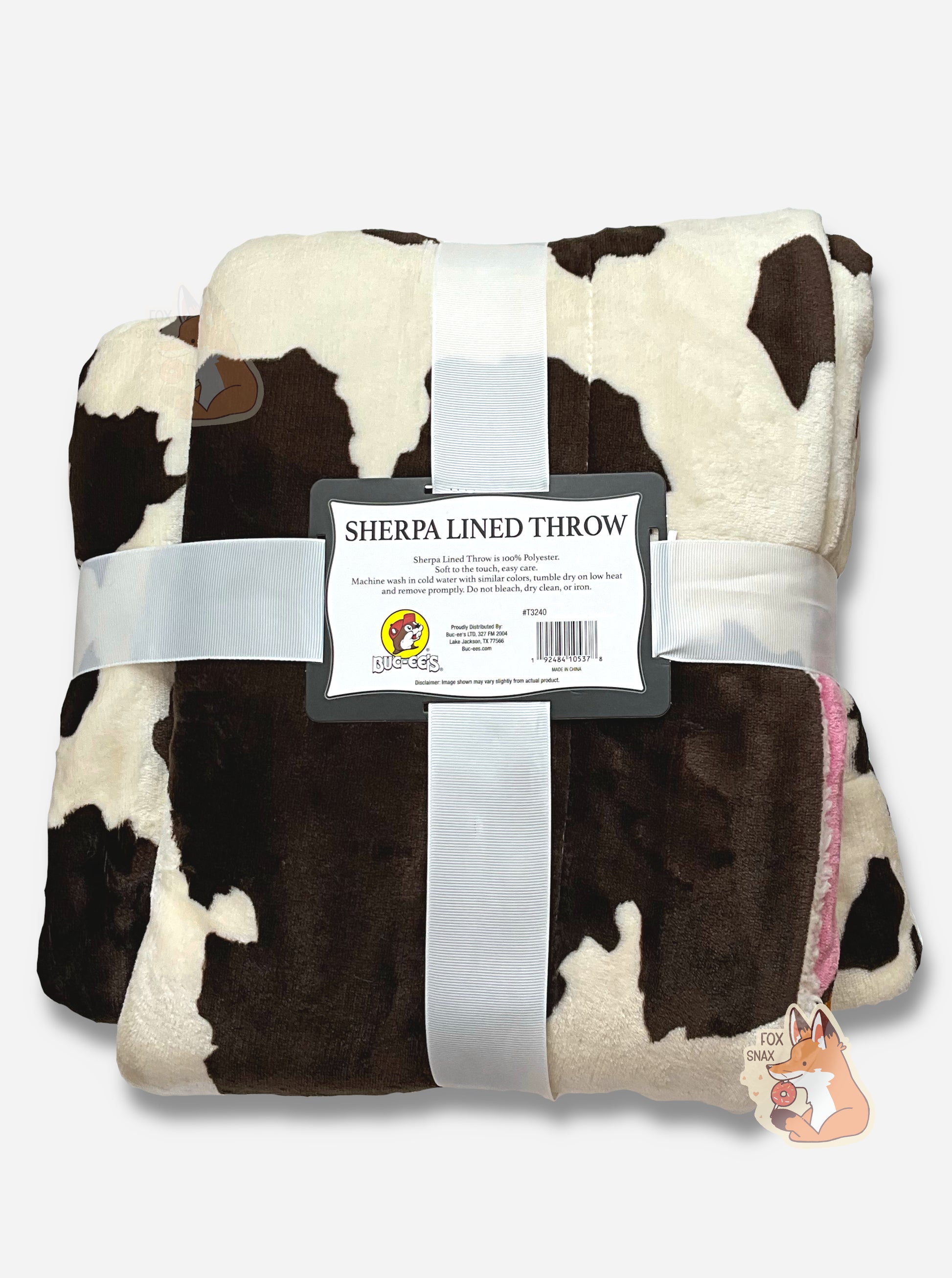 A picture of a wrapped, black and white cow-spotted fluffy blanket.  The blanket is wrapped with a translucent thick white ribbon.  In the center of the ribbon is a rectangular tag that reads SHERPA LINED THROW, then Sherpa Lined throw is 100% Polyester, Soft to the touch, easy care.  Machine wash in cold water with similar colors, tumble dry on low heat and remove promptly.  Do not bleach, dry clean, or iron.