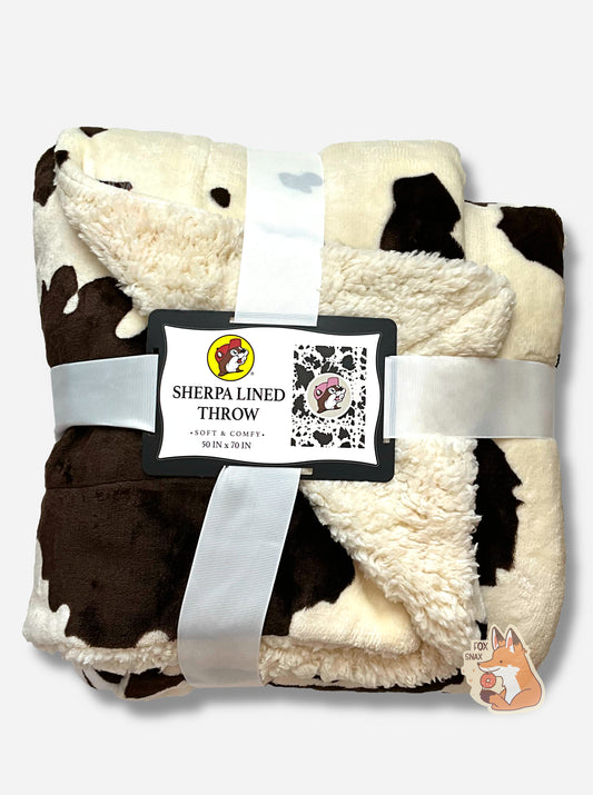 A picture of a wrapped, black and white cow-spotted fluffy blanket.  The blanket is wrapped with a translucent thick white ribbon.  In the center of the ribbon is a rectangular tag that reads SHERPA LINED THROW, SOFT& COMFY, 50 IN x 70 IN.  The center of the blanket has a circle with the Buc-ee's logo; Buc-ee's hat is light pink, and the background is light grey.
On the underside of the blanket, fluffy white sherpa lining can be seen.