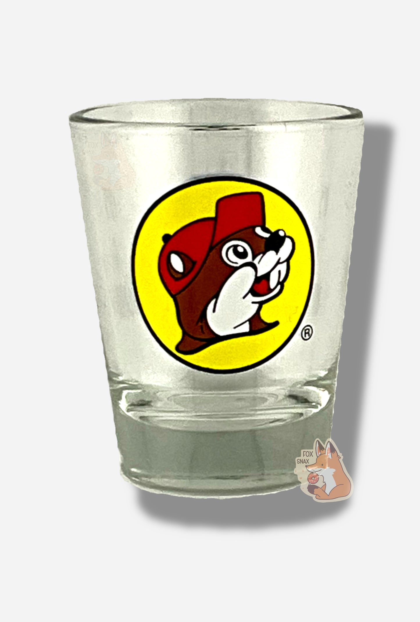 A picture of the front of glass shot glass.  The glass is clear.  In the middle of the front is the Buc-ee's logo appears inside a yellow circle; it is a cheerful beaver wearing a red ballcap, against a yellow circle background.  