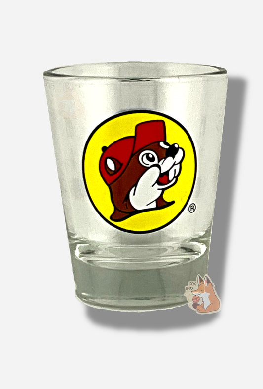 A picture of the front of glass shot glass.  The glass is clear.  In the middle of the front is the Buc-ee's logo appears inside a yellow circle; it is a cheerful beaver wearing a red ballcap, against a yellow circle background.  