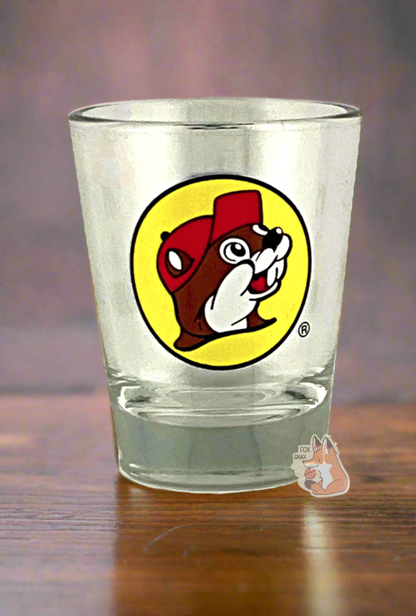 A picture of the front of glass shot glass.  The glass is clear.  In the middle of the front is the Buc-ee's logo appears inside a yellow circle; it is a cheerful beaver wearing a red ballcap, against a yellow circle background.  The shot glass is sitting on a dark wooded bar.