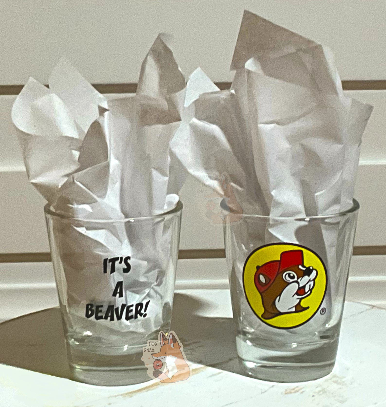 A picture of two shot glasses to demonstrate the look of the It's a Beaver Shot Glass better.  On the left, the back of the glass is displayed, which has black text with IT'S A BEAVER!.  On the right, is the front of the shot glass, with the Buc-ee The Beaver logo on it.  Both glasses have paper in them to better illustrate the text/icon.  The glasses are sitting on a white wooden bench, and a white wooden background frames them both.