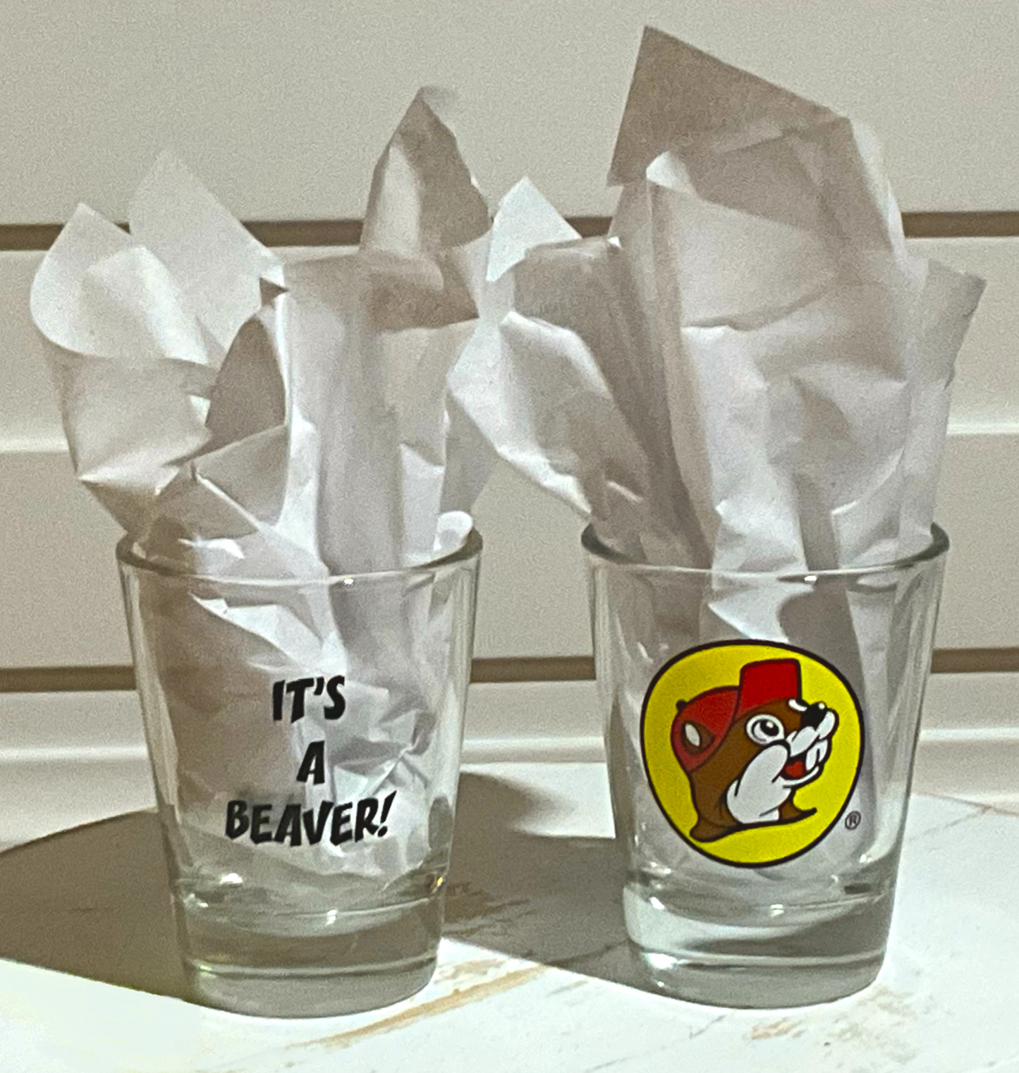 A picture of two shot glasses to demonstrate the look of the It's a Beaver Shot Glass better.  On the left, the back of the glass is displayed, which has black text with IT'S A BEAVER!.  On the right, is the front of the shot glass, with the Buc-ee The Beaver logo on it.  Both glasses have paper in them to better illustrate the text/icon.  The glasses are sitting on a white wooden bench, and a white wooden background frames them both.