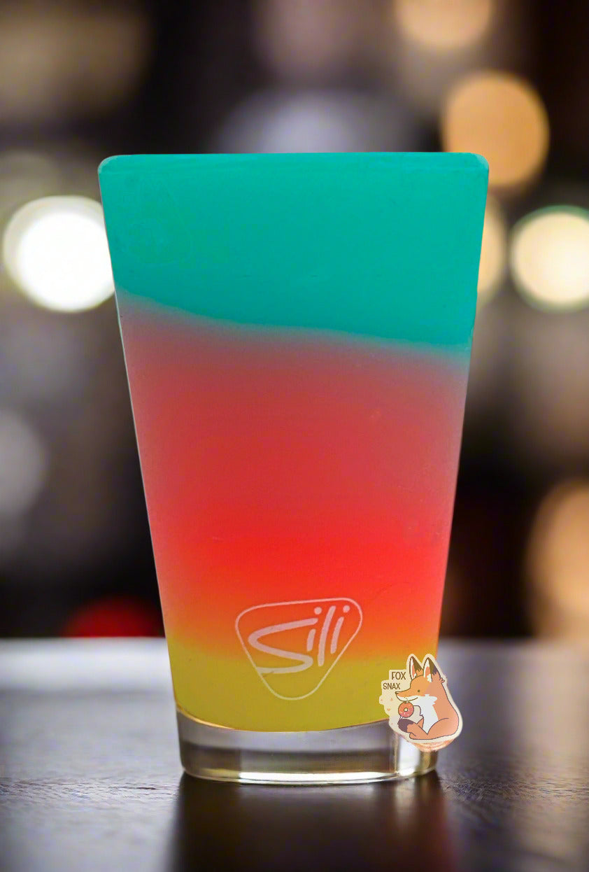 A picture of the back of a 16 oz pint glass.  The glass is made of silicone, instead of actual glass, and starts with a blue/green gradient at the top, which fades to purple, then red in the middle, which fades to orange, then yellow at the bottom.  The bottom of the glass has a logo in white which reads SILI. The glass is sitting on a wooden bar; blurred bottles can be seen against the back of the bar.