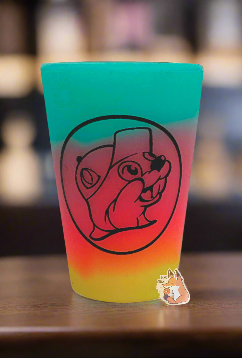 A picture of shot glass pint glass.  The glass is made of silicone, instead of actual glass, and starts with a blue/green gradient at the top, which fades to purple, then red in the middle, which fades to orange, then yellow at the bottom.  The front of the pint glass has, in a black outline, the Buc-ee's logo - a cheerful beaver wearing a ball cap.  The glass is sitting on a wooden bar; blurred bottles can be seen against the back of the bar.