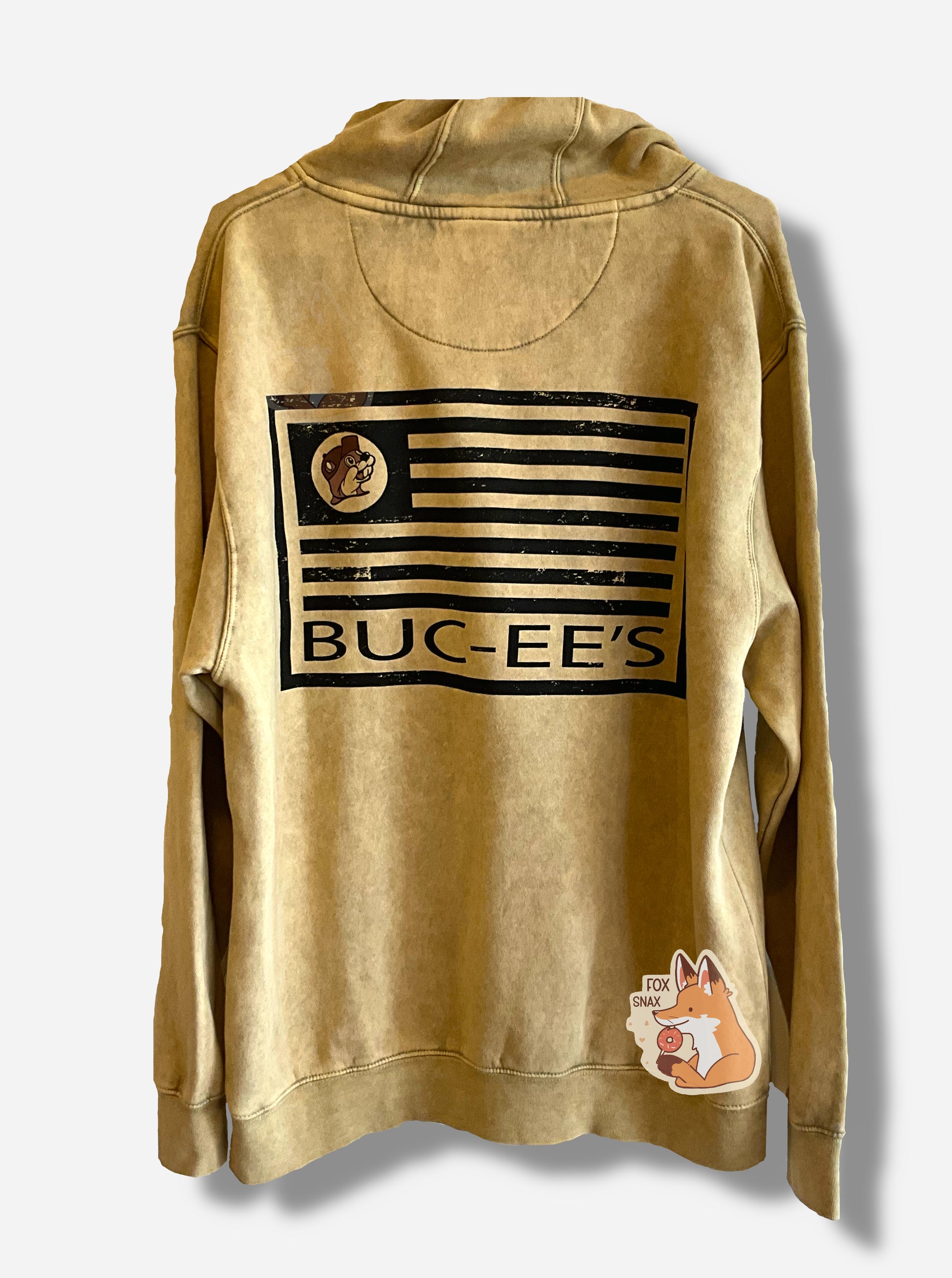 A picture of the back of a large, tan hoodie sweatshirt.  The back shows an outline of an American flag, but the stripes are all in black, and instead of stars, the same tan logo as the front of the iconic beaver is shown.  Beneath the flag, all in black text, read the words BUC-EE'S.