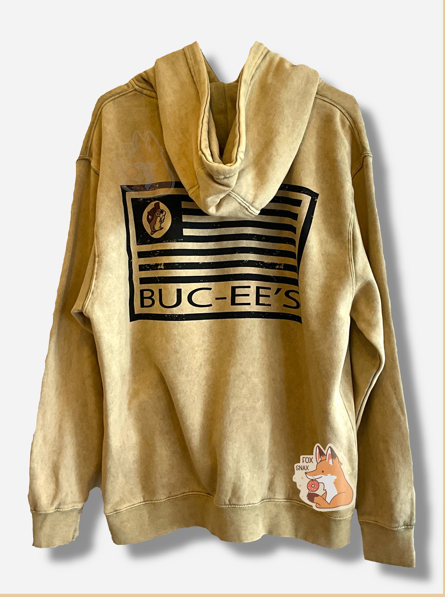 A picture of the back of a large, tan hoodie sweatshirt.  The back shows an outline of an American flag, but the stripes are all in black, and instead of stars, the same tan logo as the front of the iconic beaver is shown.  Beneath the flag, all in black text, read the words BUC-EE'S.  In this picture, the tan hood is draped over the flag on the back.