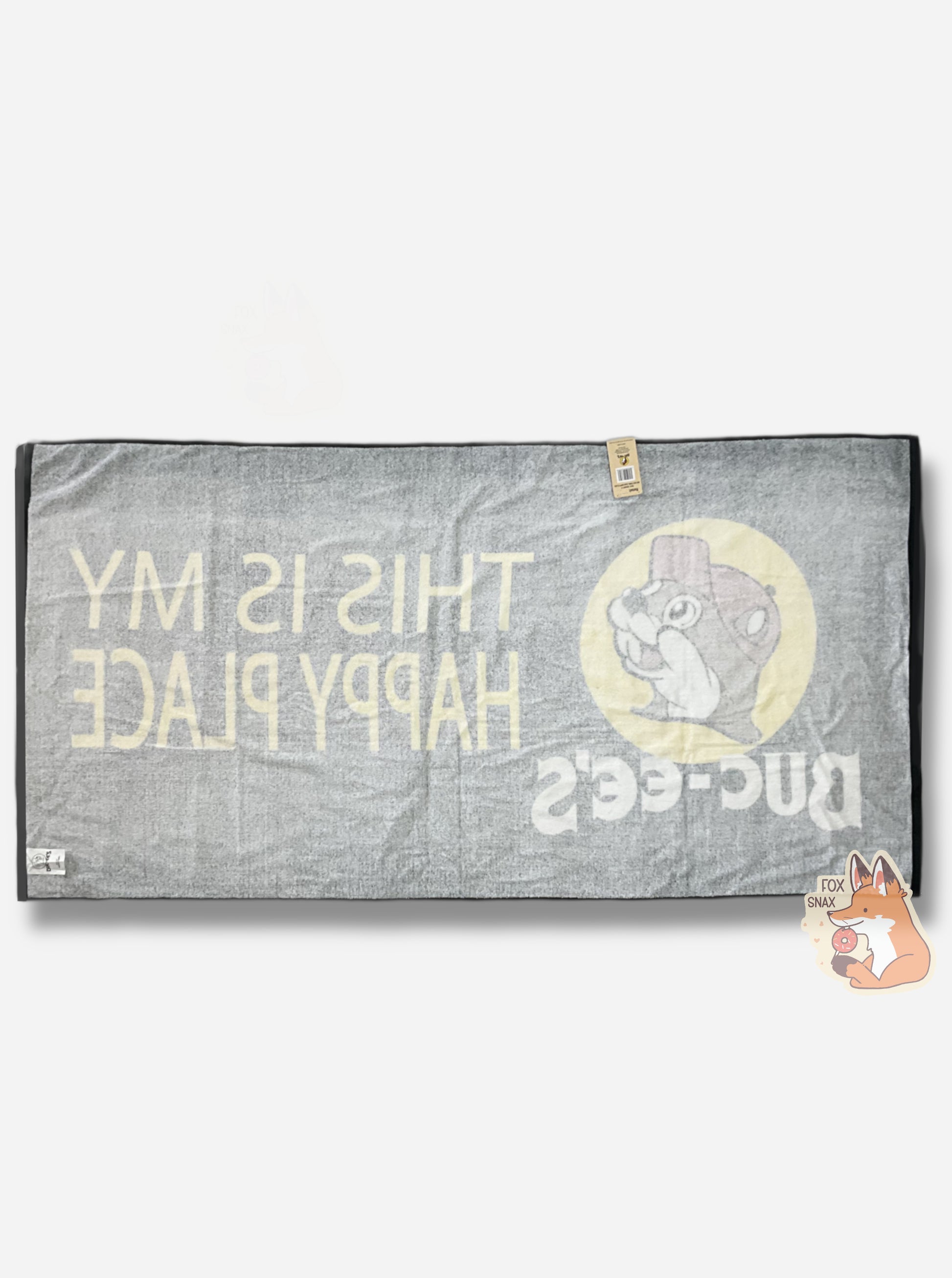 A picture of the back of an  unwrapped, 32x64, fluffy black beach towel.  The back of the towel in white with a black border.  Faintly the front of the towel can be seen; it's (backwards) and adorned with a single Buc-ee The Beaver logo on the middle left.   Buc-ee is framed by a yellow circle, and is wearing a red ball cap. Underneath the logo reads in white text, BUC-EES.  On the right side of the towel, in yellow text, reads THIS IS MY HAPPY PLACE.